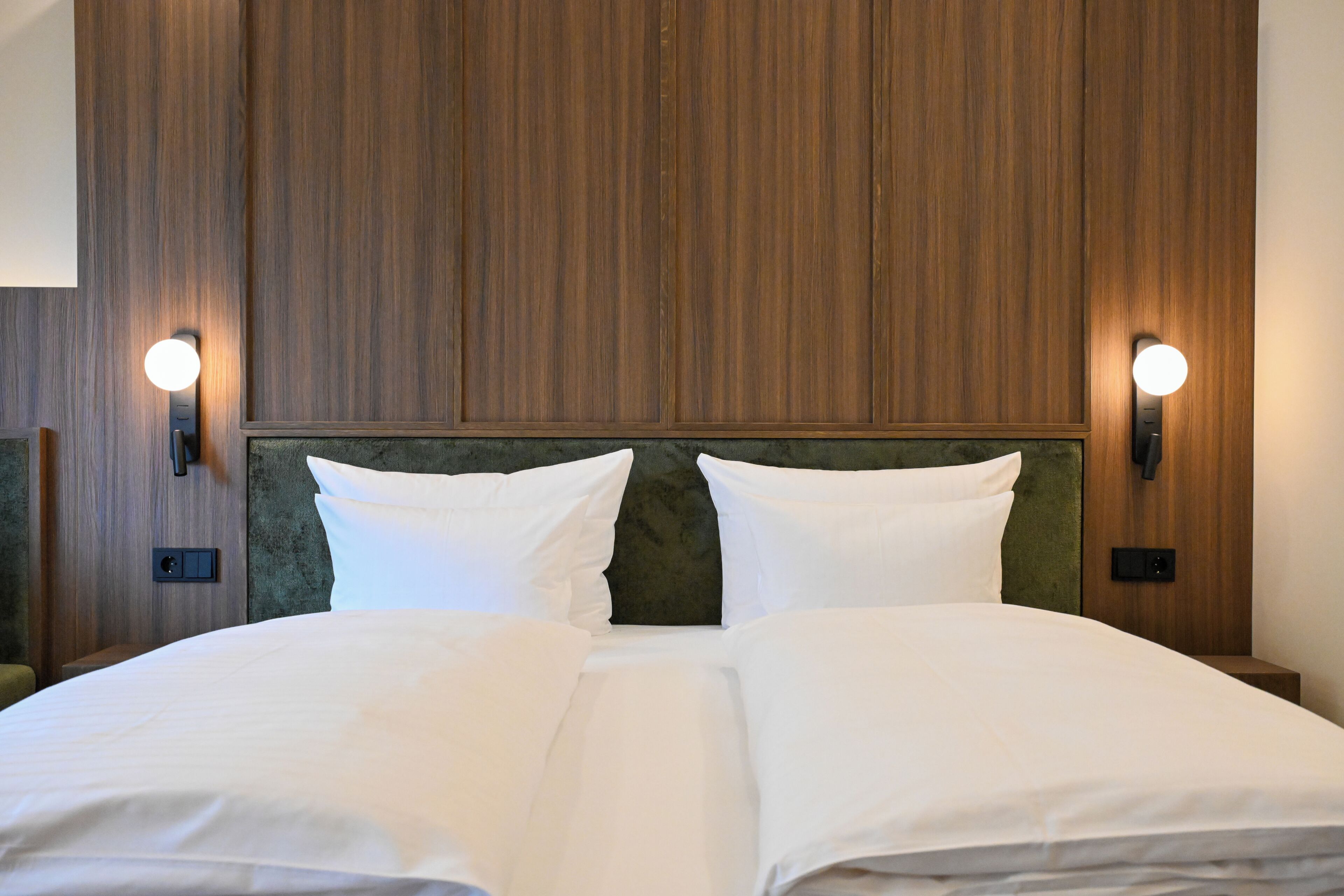 Une chambre d’hôtel avec une tête de lit en bois, deux lampes de chevet et un lit soigneusement fait avec du linge de lit blanc.