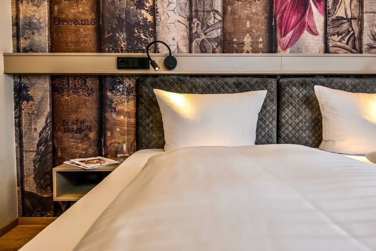 A neatly made bed with a headboard, a bedside table with a magazine, and a wall with patterned fabric and a clock.