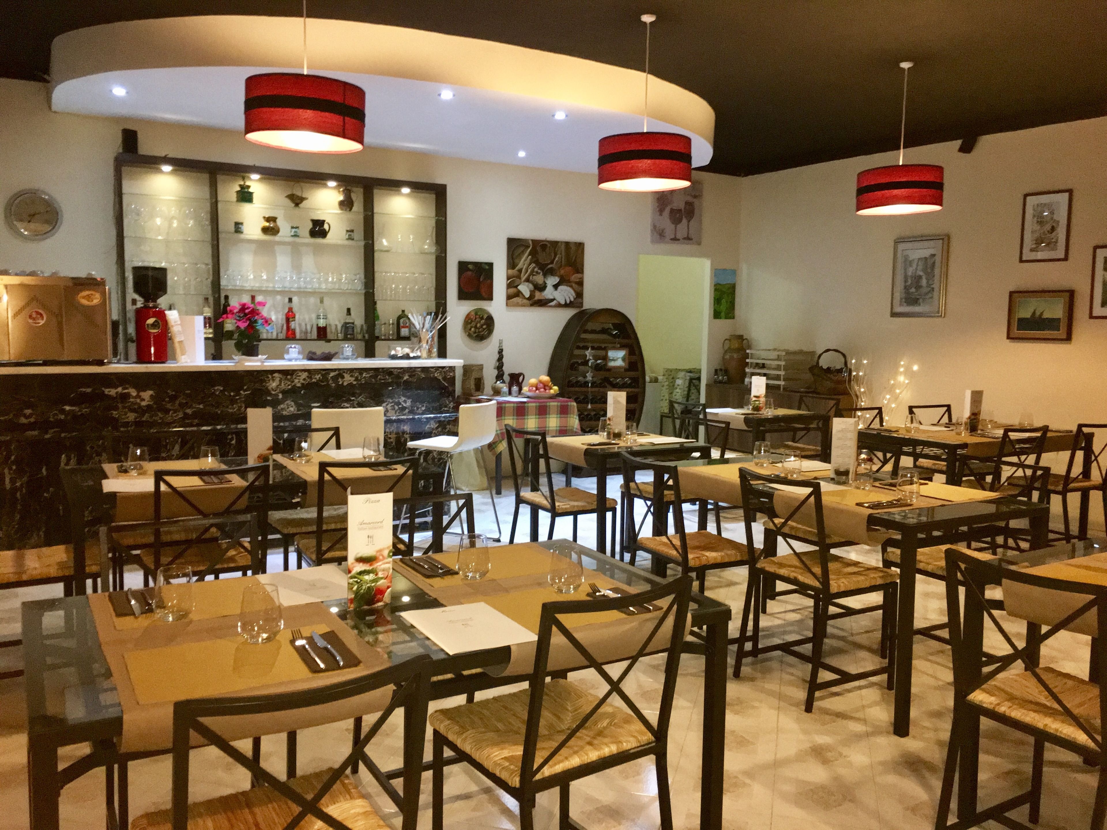 Un restaurante con varias mesas y sillas, una zona de bar con estanterías llenas de botellas y lámparas rojas colgantes.