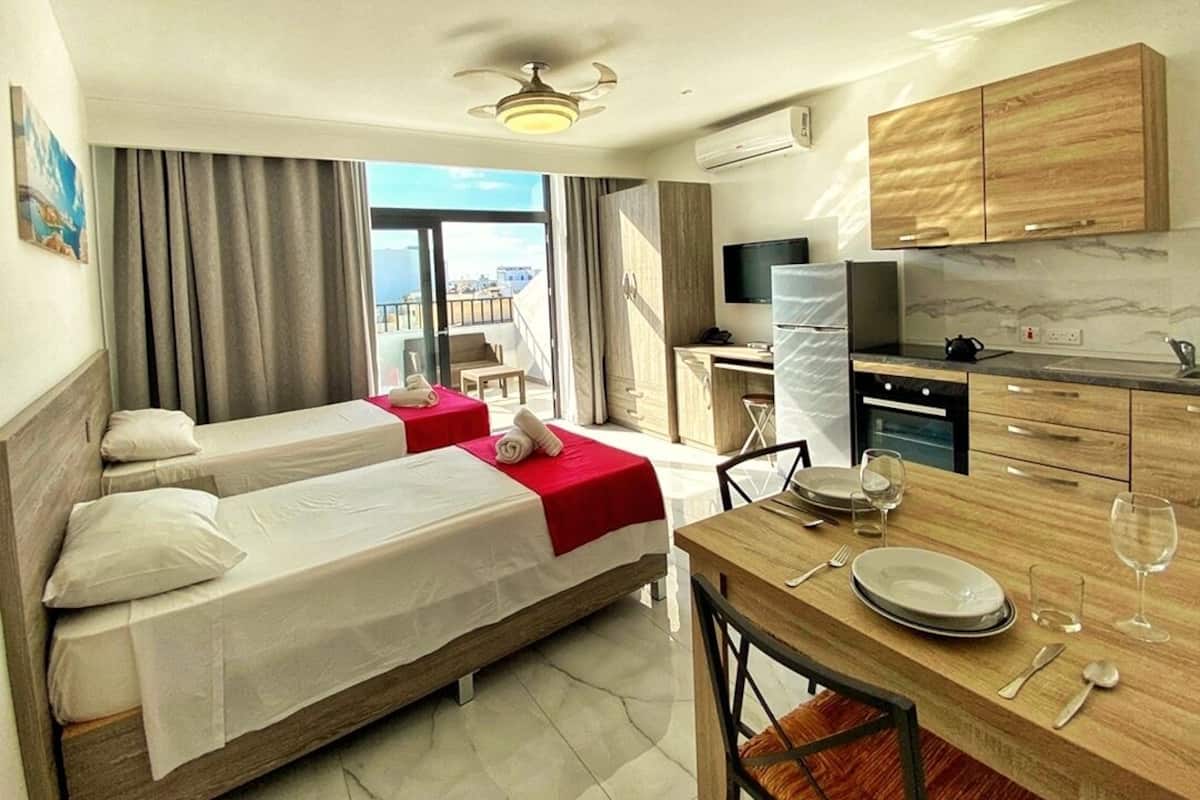 Quarto de hotel com duas camas, área de jantar, cozinha e uma grande janela com vista para a cidade.