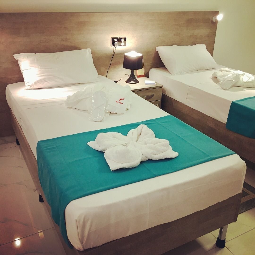 A hotel room with two beds, each with white linens and a teal bedspread. There is a nightstand with a lamp between the beds, and a pair of white towels on the beds.