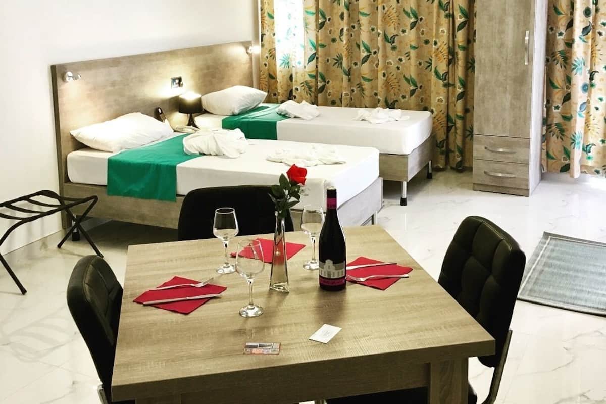 Quarto de hotel com duas camas, uma mesa de jantar com cadeiras, minibar e uma janela grande com cortinas.