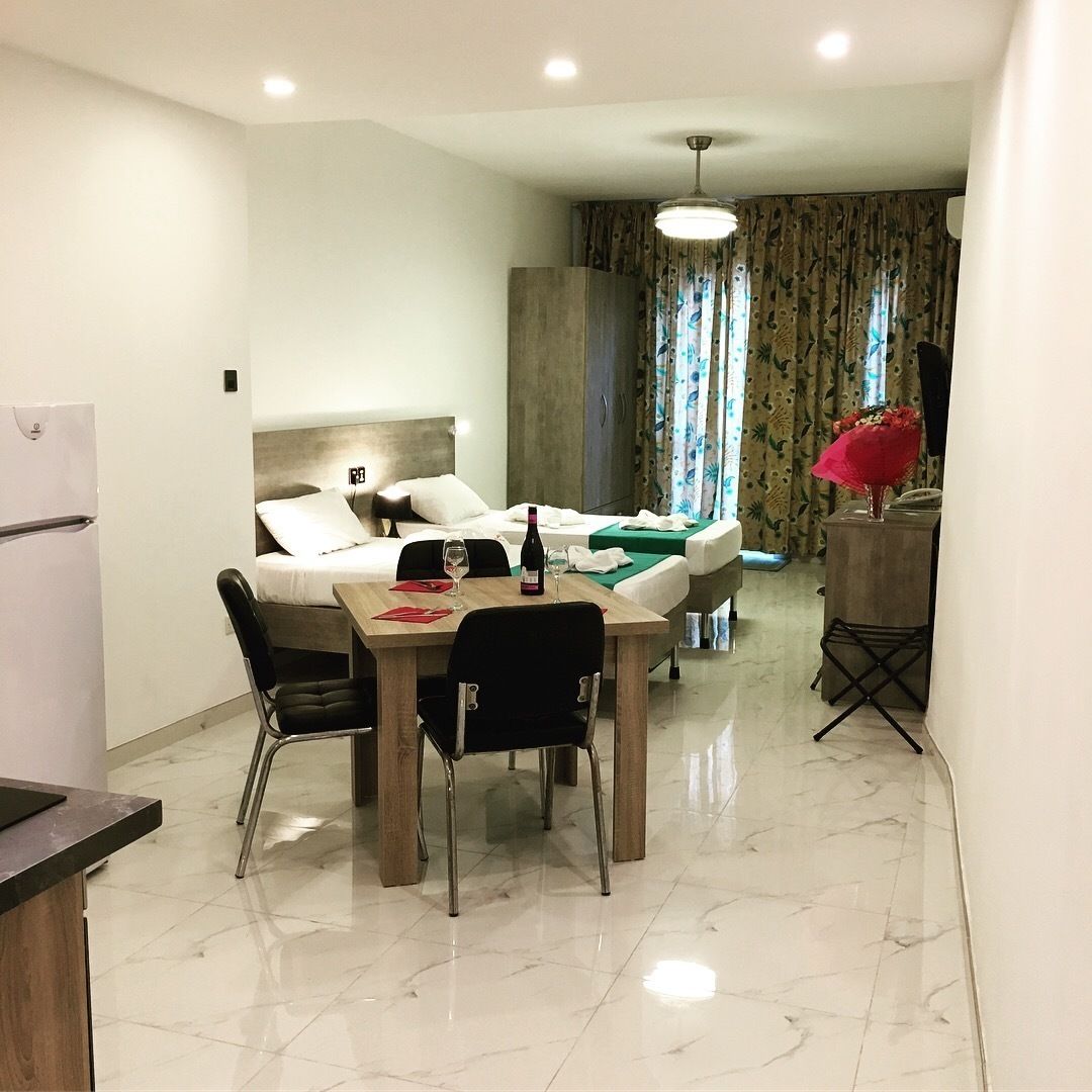 A compact hotel room with a dining area, a bed with a headboard, a small table with chairs, a refrigerator, and a window with curtains.