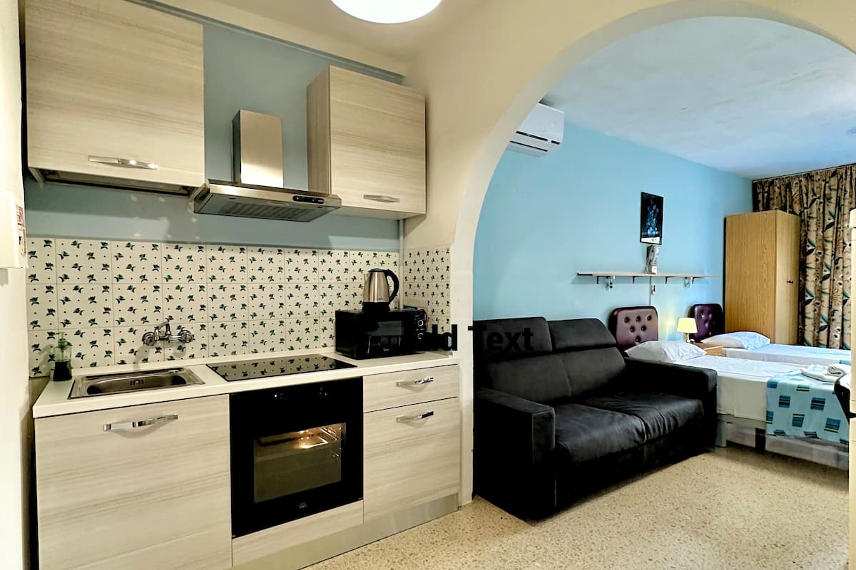 Uma cozinha compacta com forno embutido, pia e cafeteira. Há um sofá preto e uma cama com colcha estampada em azul e branco. O quarto tem parede azul clara e uma pequena janela com cortinas.