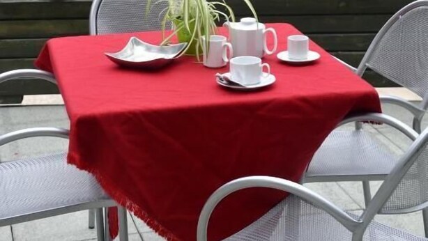 A table set for two with a red tablecloth, white chairs, and a view of a mountain and buildings in the background. - Martinella (Folgaria)