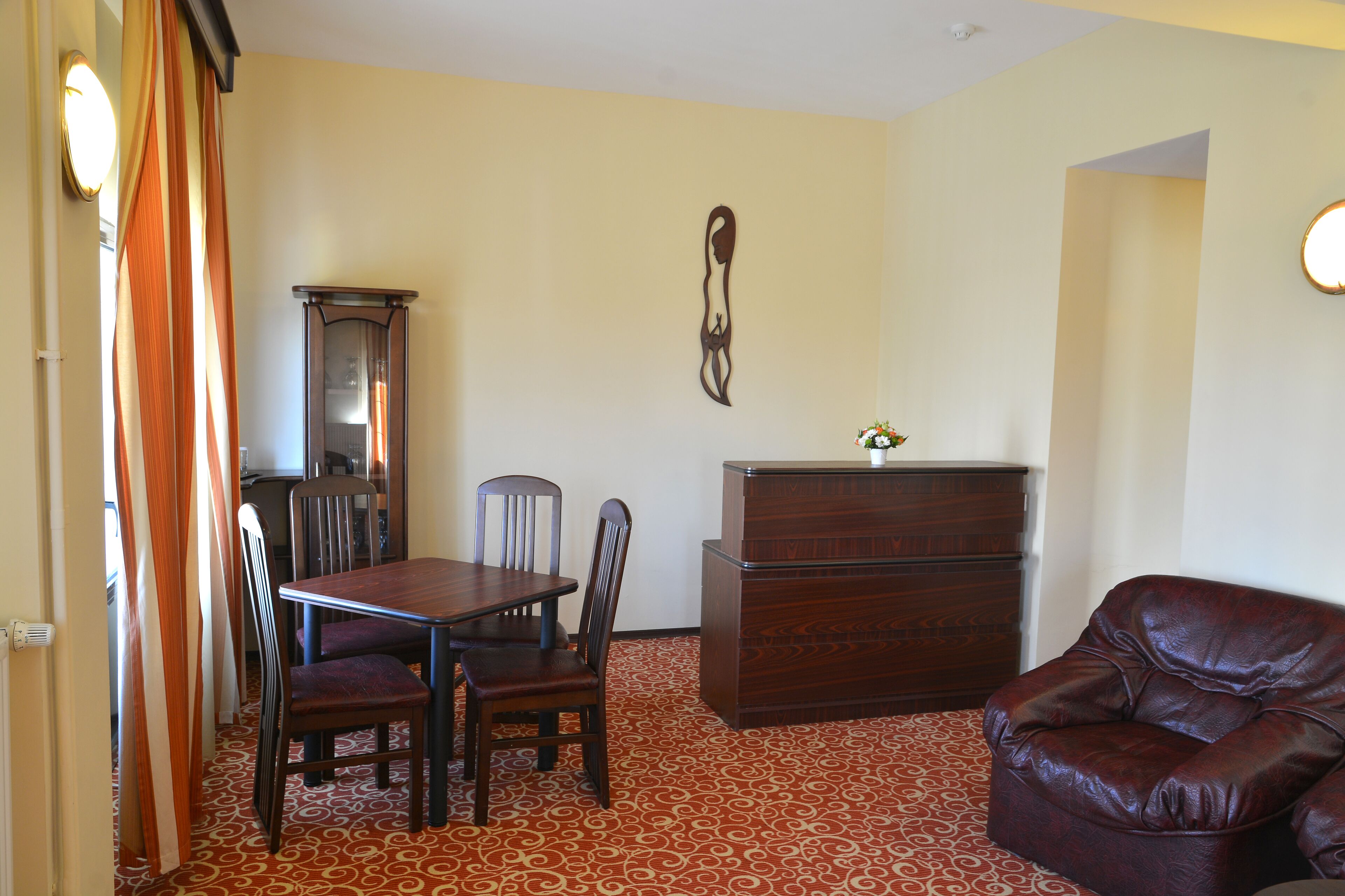 A room with a dining table, chairs, a wooden cabinet, a leather armchair, and a patterned carpet.