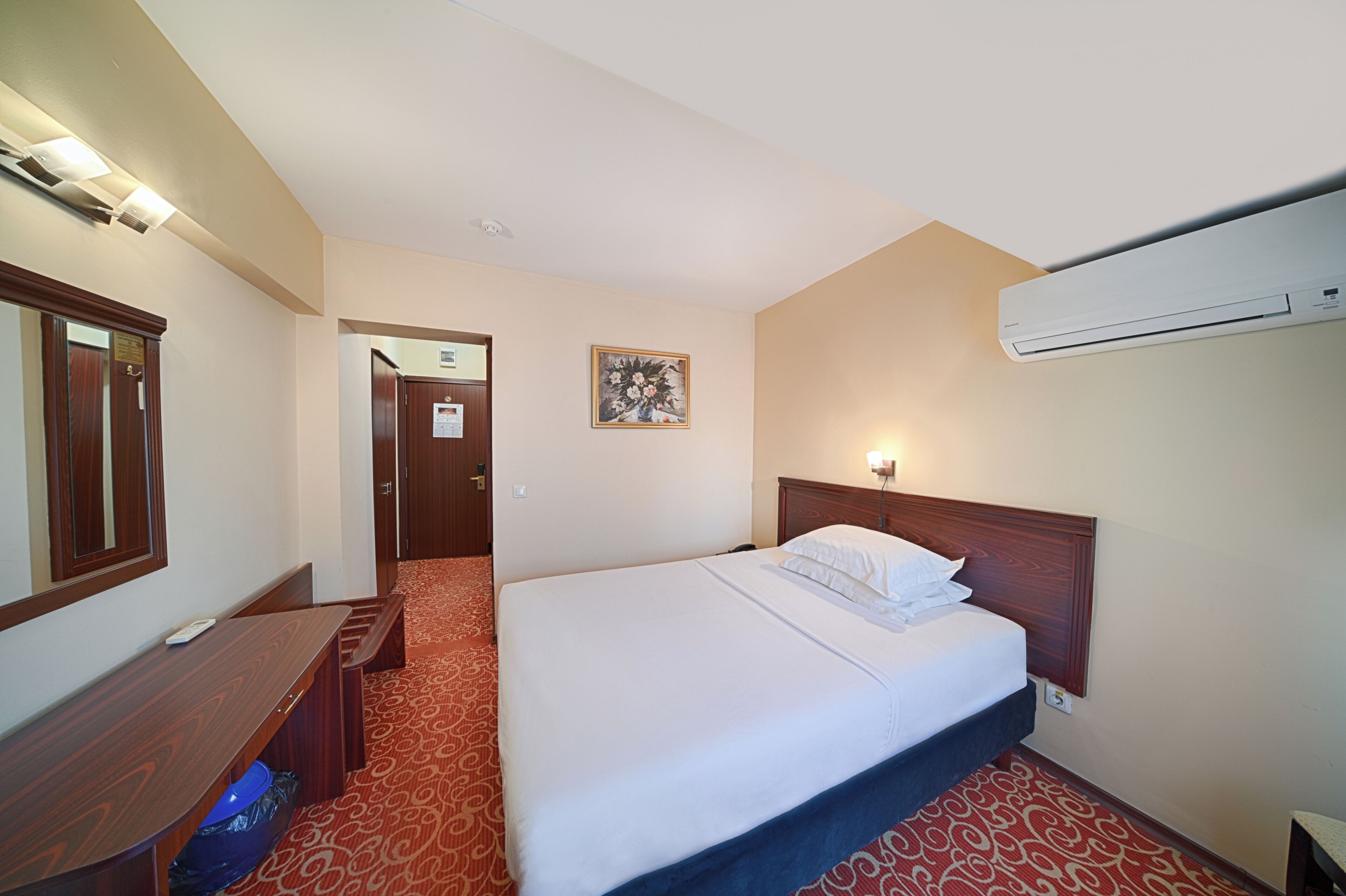 A hotel room with a bed, a desk, a mirror, a door, and an air conditioning unit.