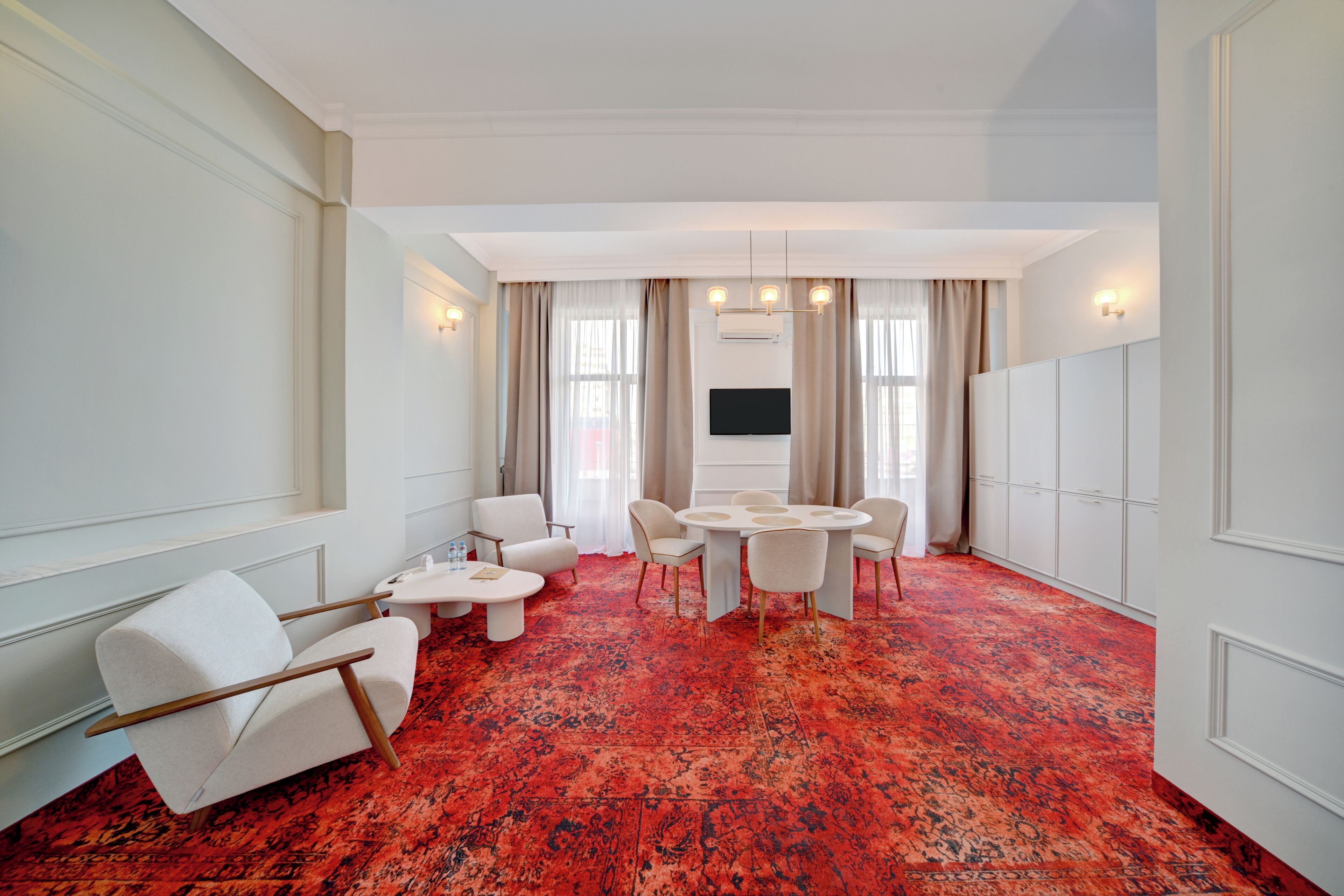 Habitación de hotel con una alfombra roja de estampado grande, una mesa redonda de comedor con sillas, un televisor de pantalla plana montado en la pared y un rincón de estar con dos sillones y una mesita.