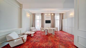 Habitación de hotel con una alfombra roja de estampado grande, una mesa redonda de comedor con sillas, un televisor de pantalla plana montado en la pared y un rincón de estar con dos sillones y una mesita.