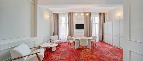 Une chambre d’hôtel dotée d’un grand tapis rouge à motifs, d’une table de salle à manger ronde avec des chaises, d’un téléviseur à écran plat fixé au mur et d’un coin salon avec deux fauteuils et une petite table.