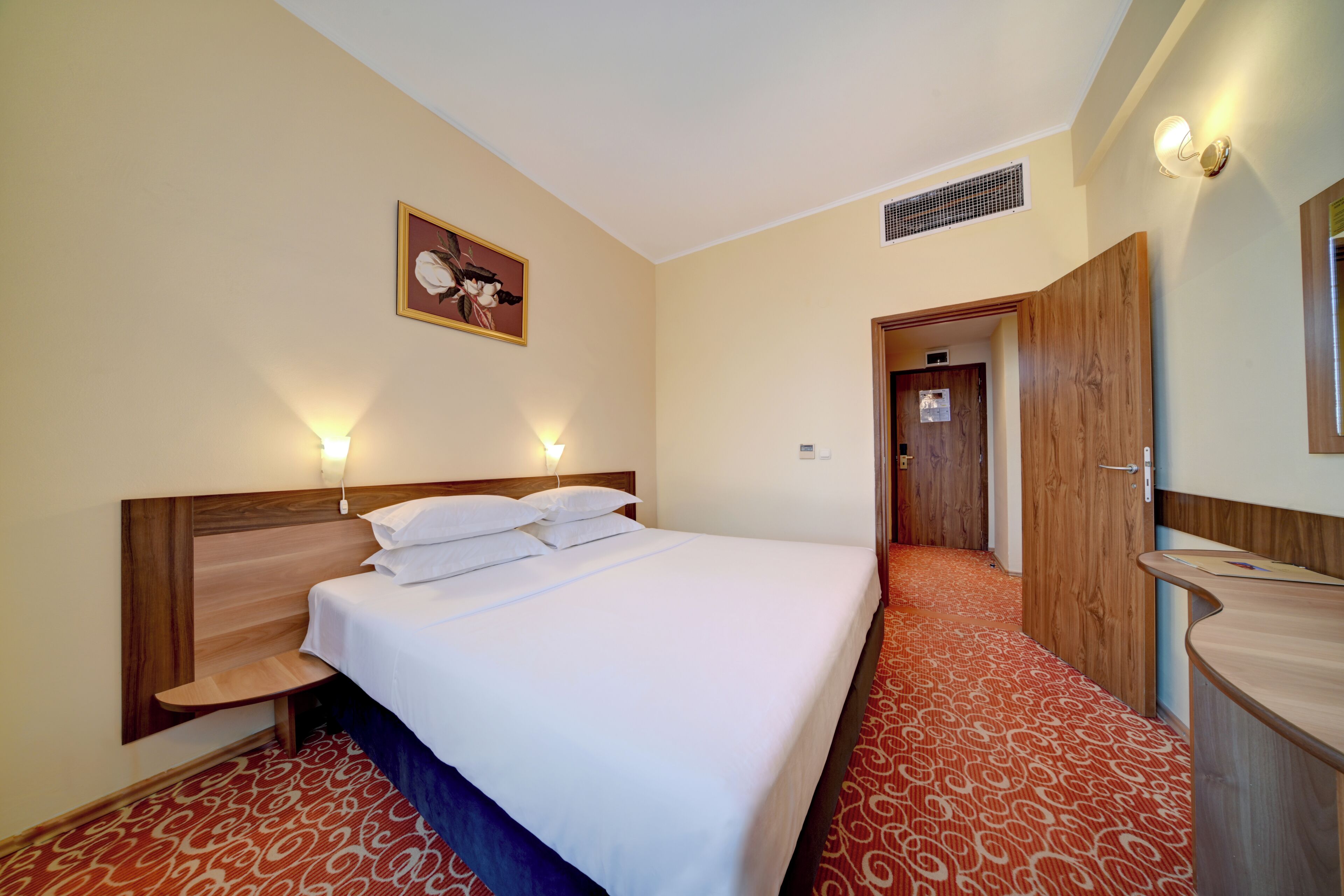 Ein Hotelzimmer mit einem Bett, Nachttischlampen, einem hölzernen Kopfteil, einem roten Teppich mit Muster sowie einer Holztür und einem Spiegel.