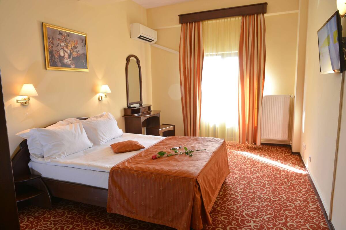 A hotel room with a bed, bedside lamps, a mirror, a window with curtains, and a radiator.