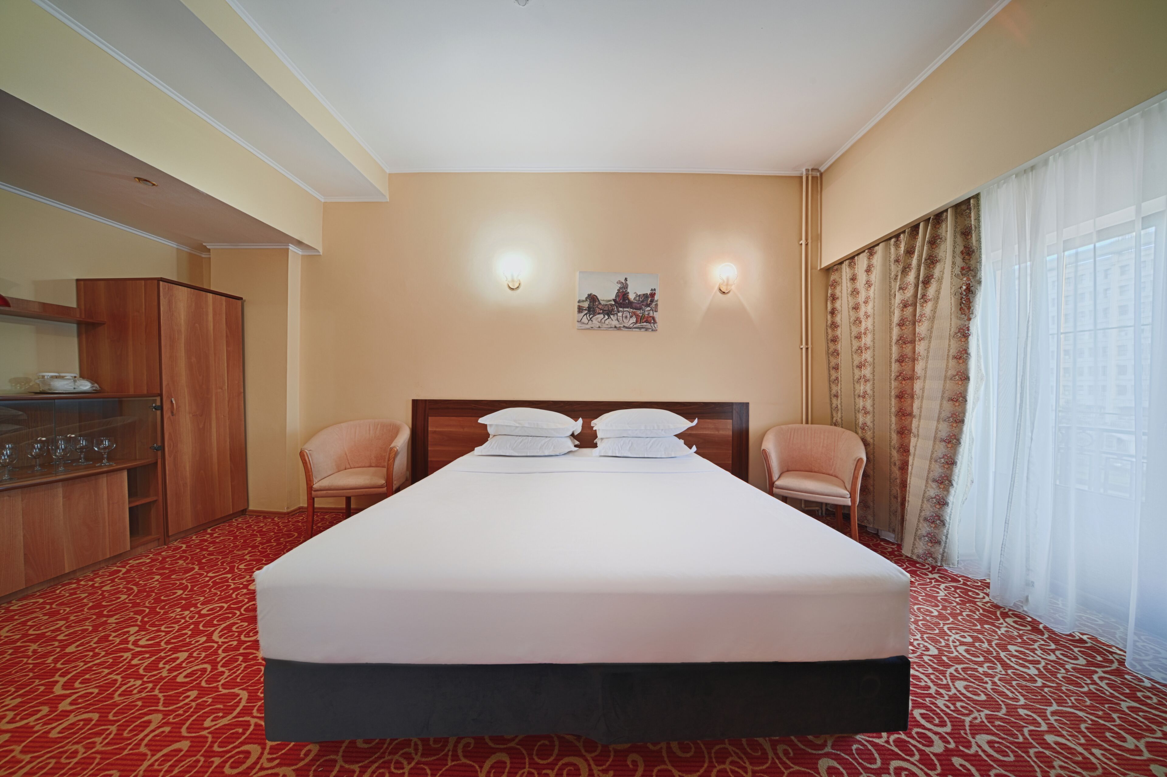 Quarto de hotel com uma cama grande, duas cadeiras, um guarda-roupa de madeira e um quadro na parede.