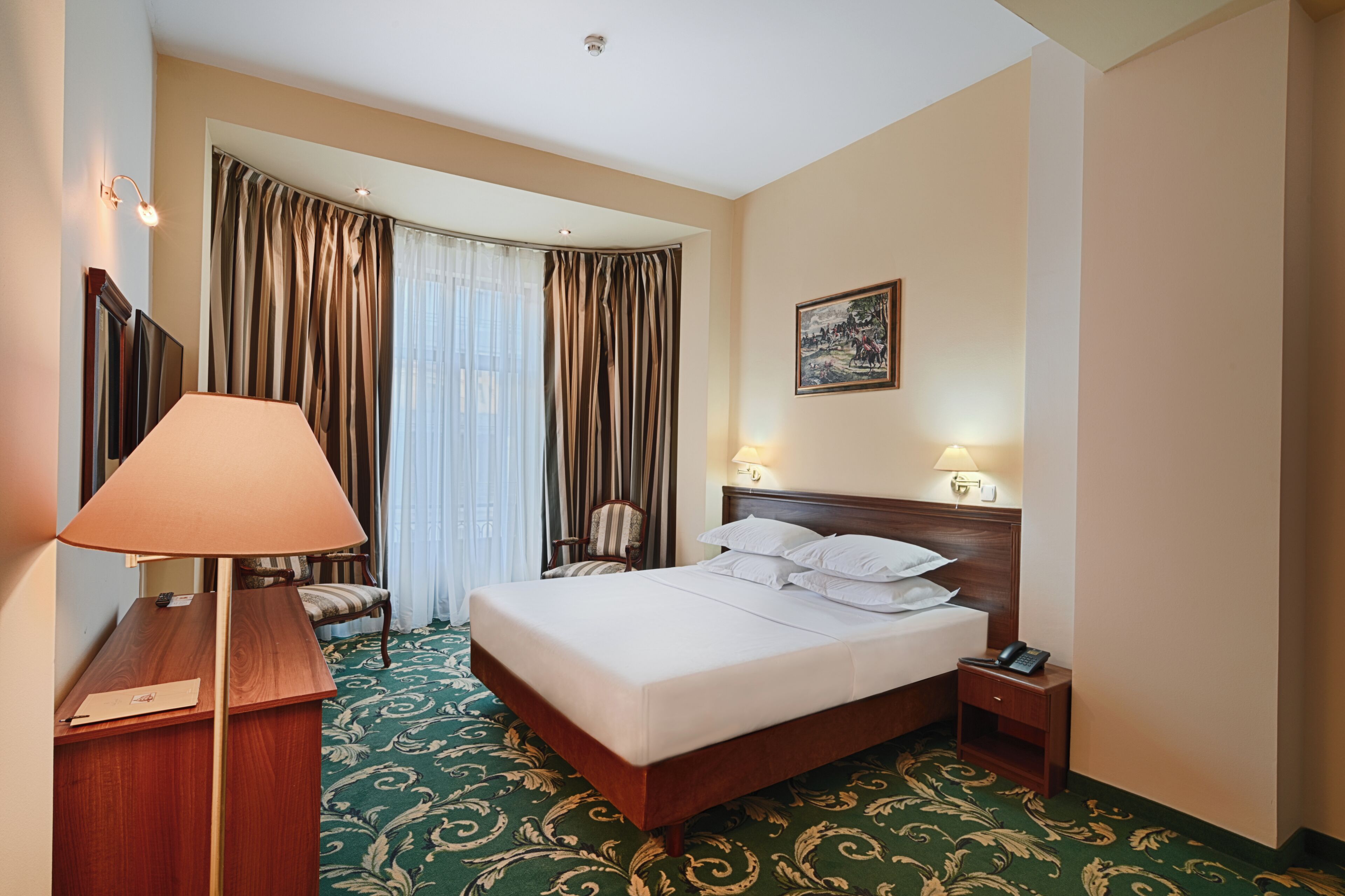 Habitación de hotel con cama, escritorio, lámpara, silla, un cuadro en la pared y una ventana con cortinas.