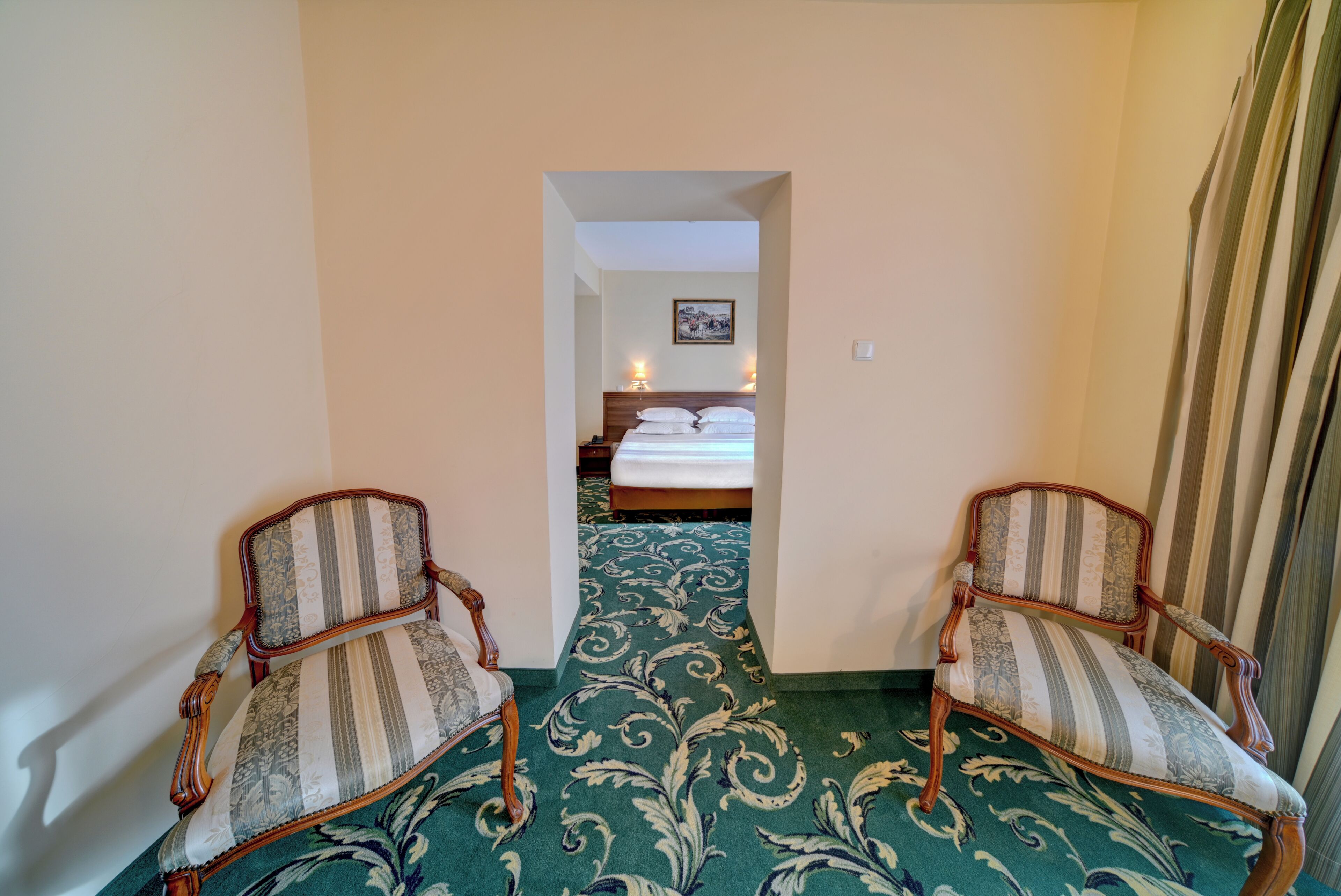 A hallway leading to a bedroom with a bed, two upholstered chairs, and a patterned carpet.