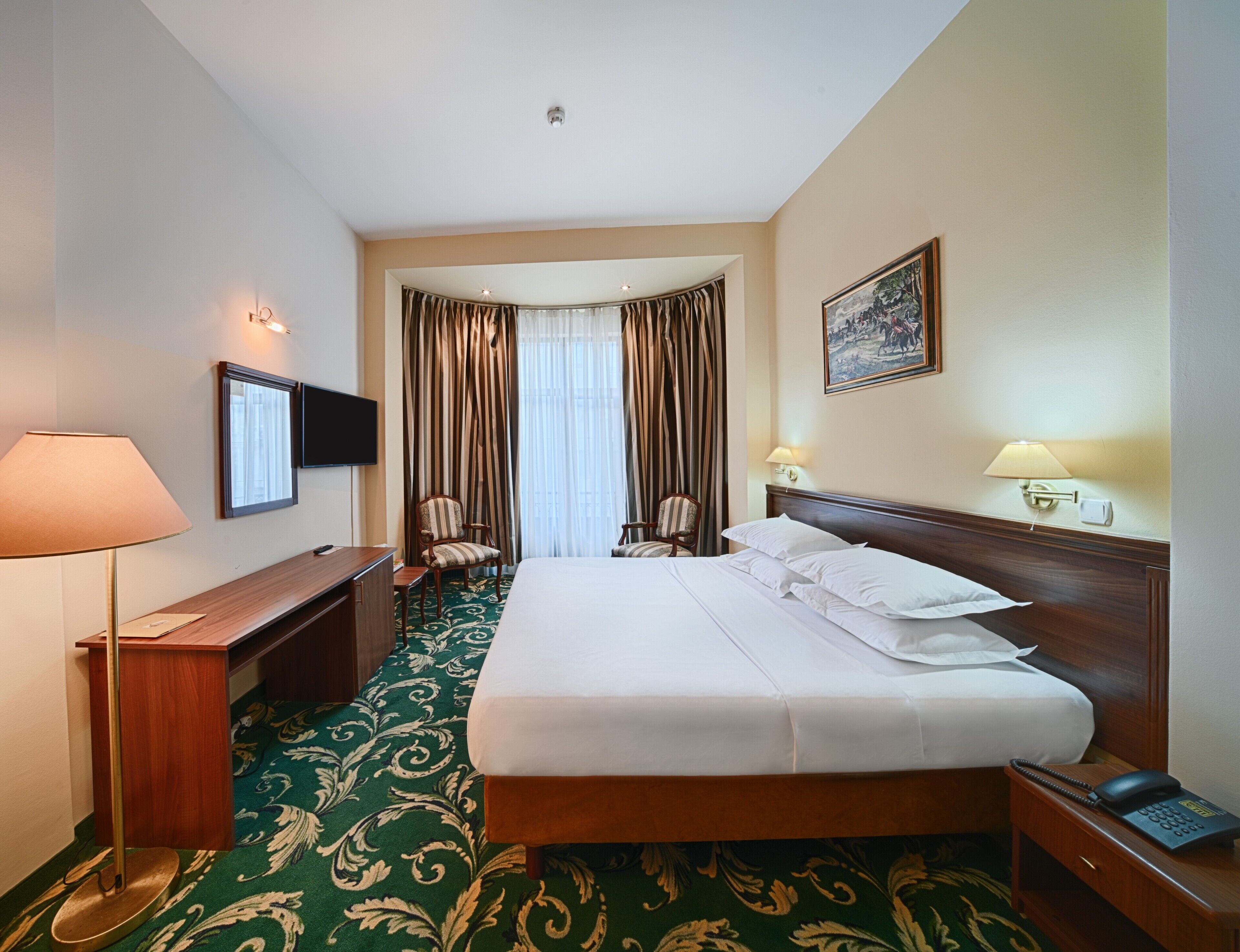 A hotel room with a large bed, a desk with a lamp, a chair, a TV, and a painting on the wall.