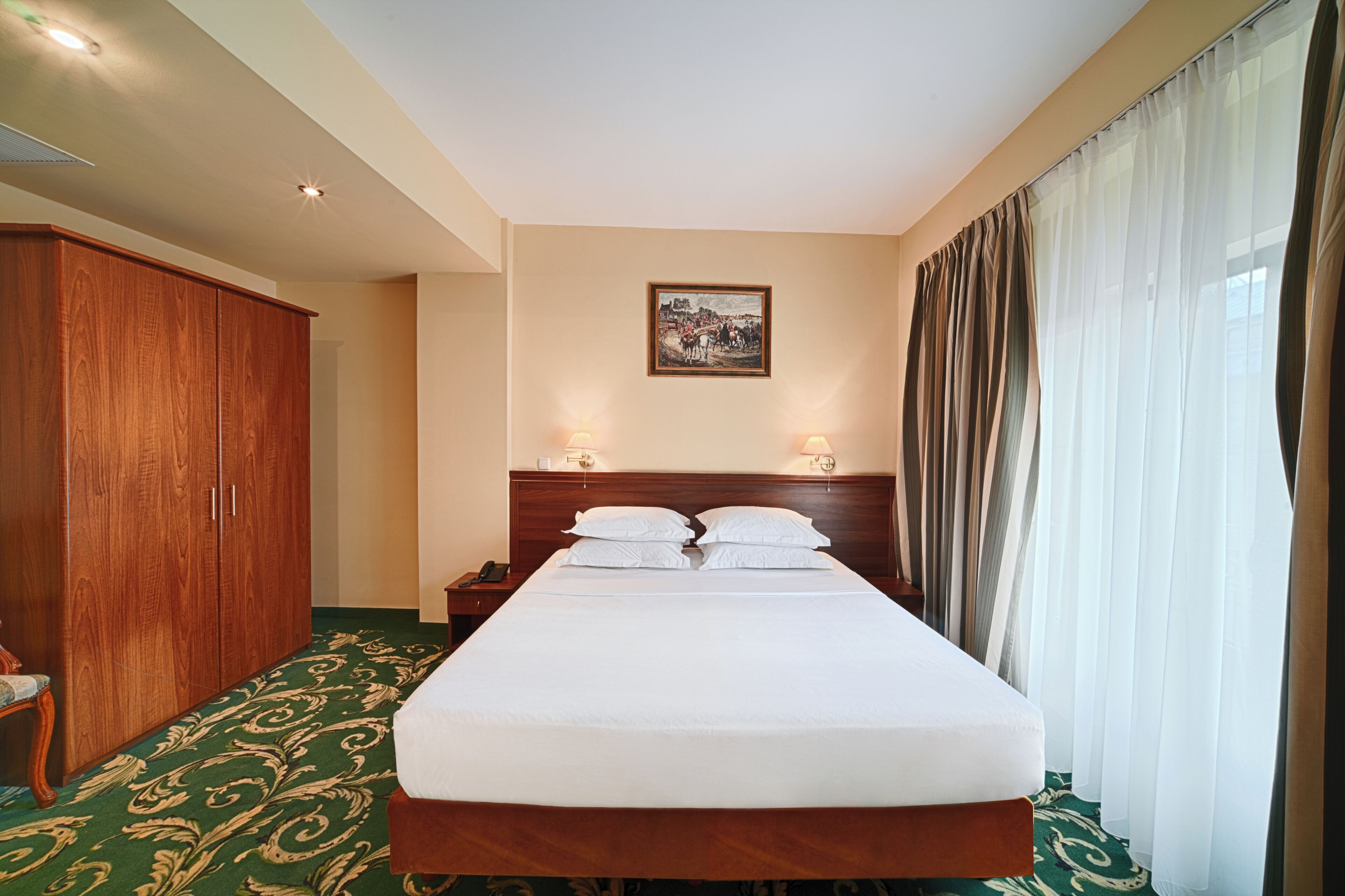 A hotel room with a large bed, wooden wardrobe, two bedside lamps, a framed picture on the wall, and a window with curtains.