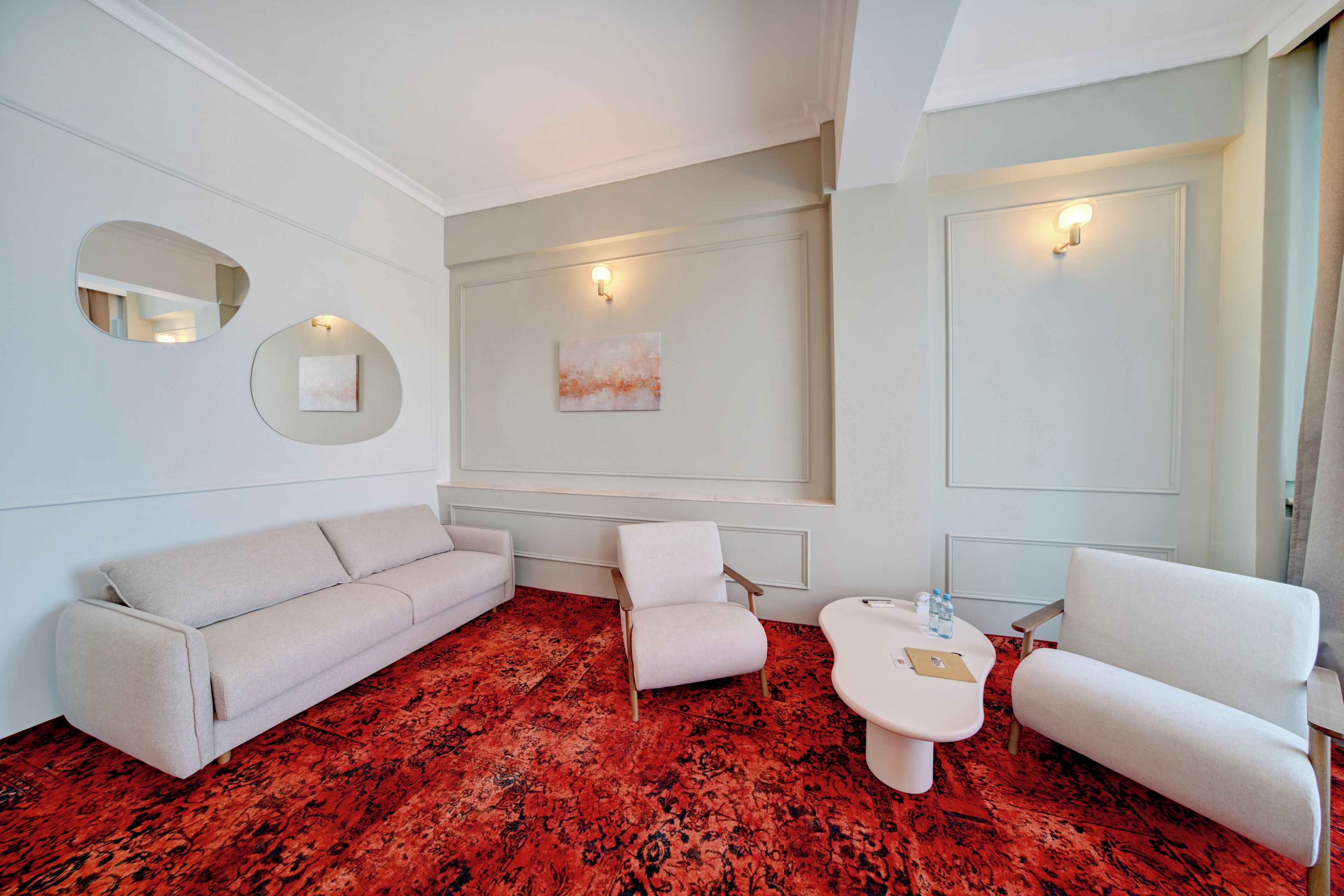A modern room with a red patterned carpet, a white sofa, a white armchair, a small white table, and a wall-mounted light fixture.