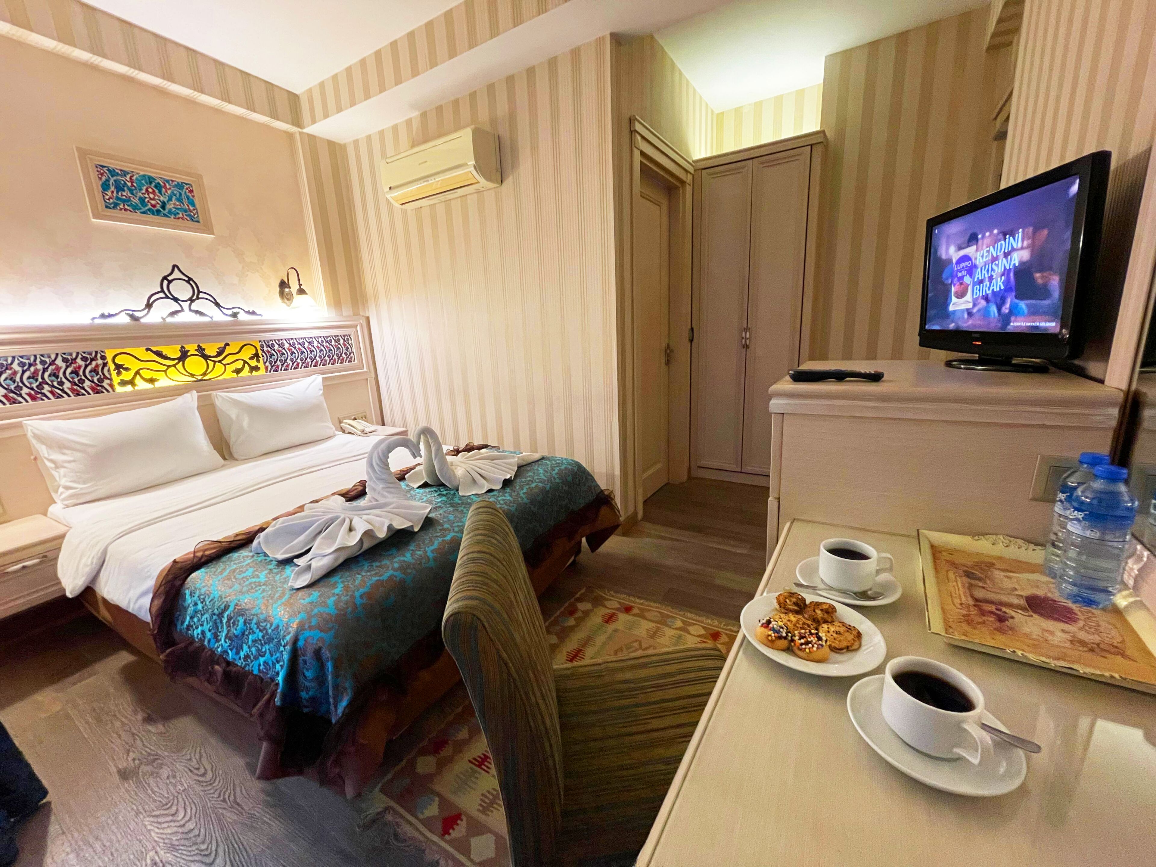 A hotel room with a bed, a desk with a coffee cup and pastries, a television, and an air conditioning unit.