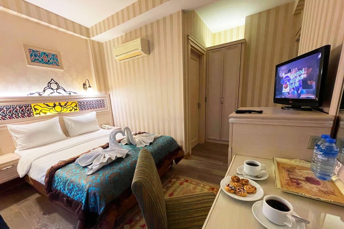 A hotel room with a bed, a desk with a coffee cup and pastries, a television, and an air conditioning unit.