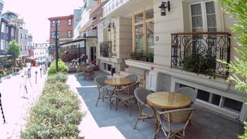 An outdoor cafe area with wicker chairs and tables, surrounded by greenery and a street view.