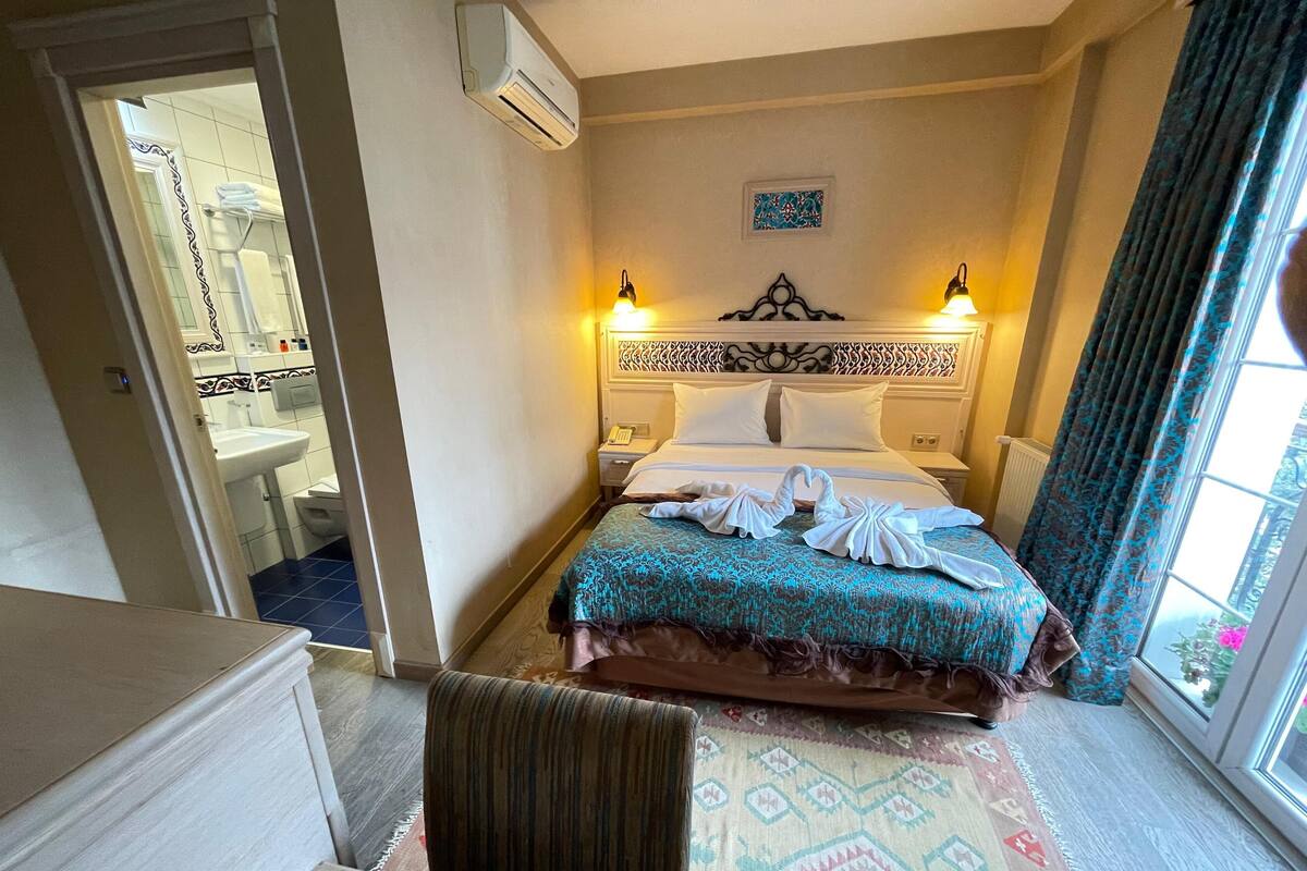 A hotel room with a bed, bedside lamps, a headboard, a window with curtains, a bathroom visible through an open door, and a patterned rug.