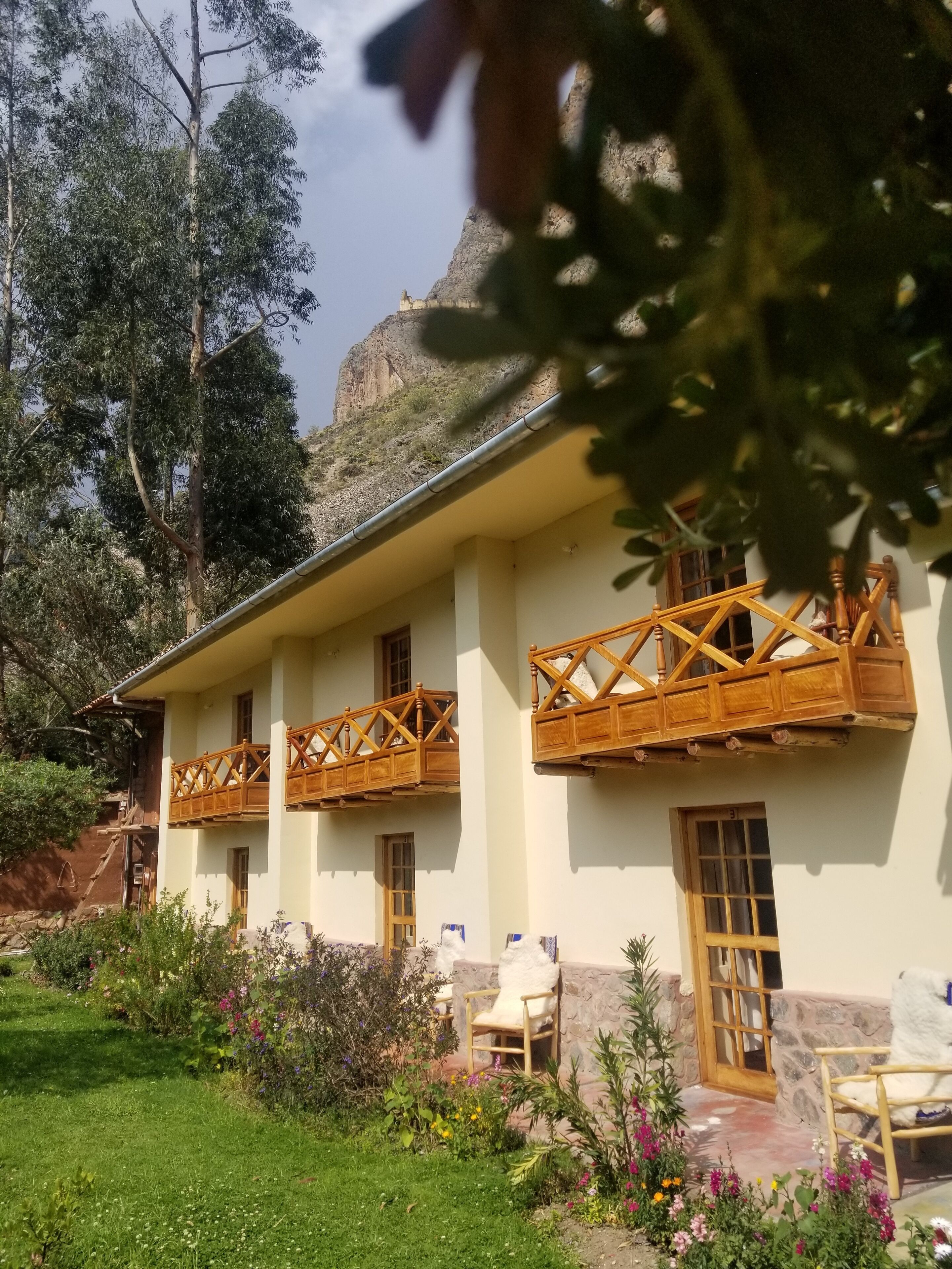 Un edificio de dos plantas con balcones de madera, una entrada de piedra y un jardín con diversas plantas y flores.