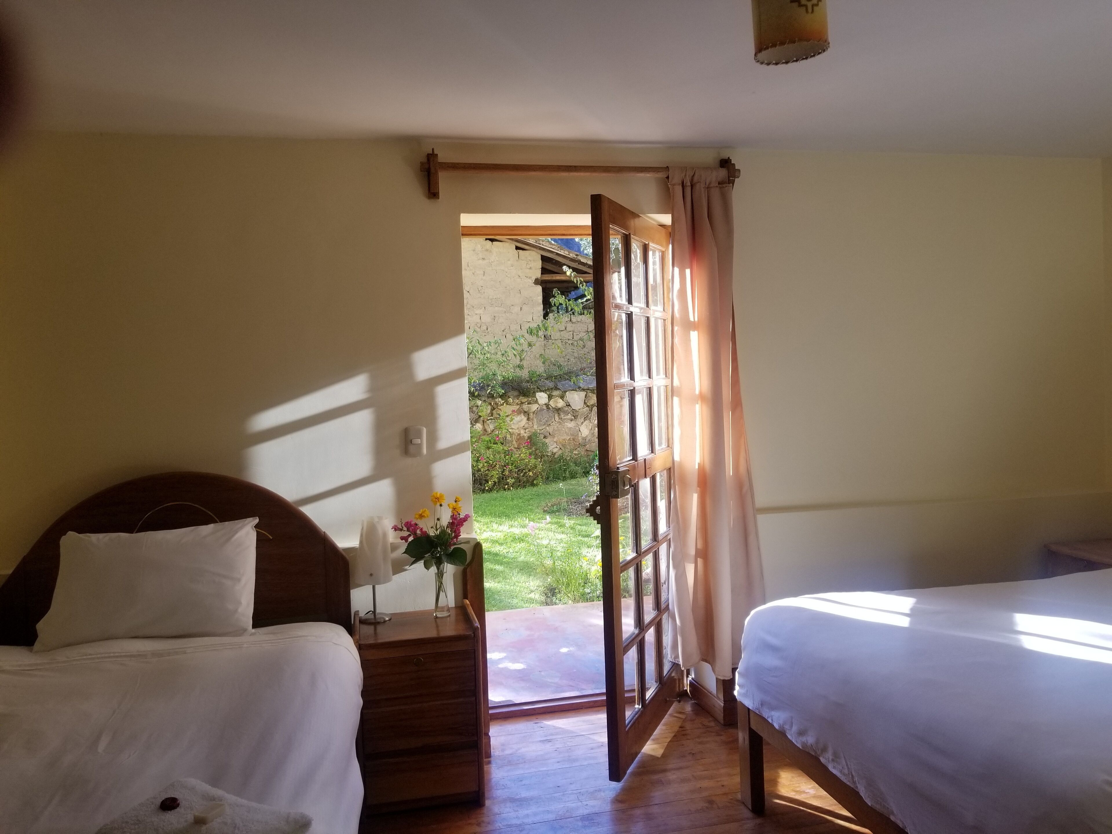 Ein Schlafzimmer mit zwei Betten, einem Nachttisch mit Blumenvase und Blick auf einen Garten durch eine offene Tür.