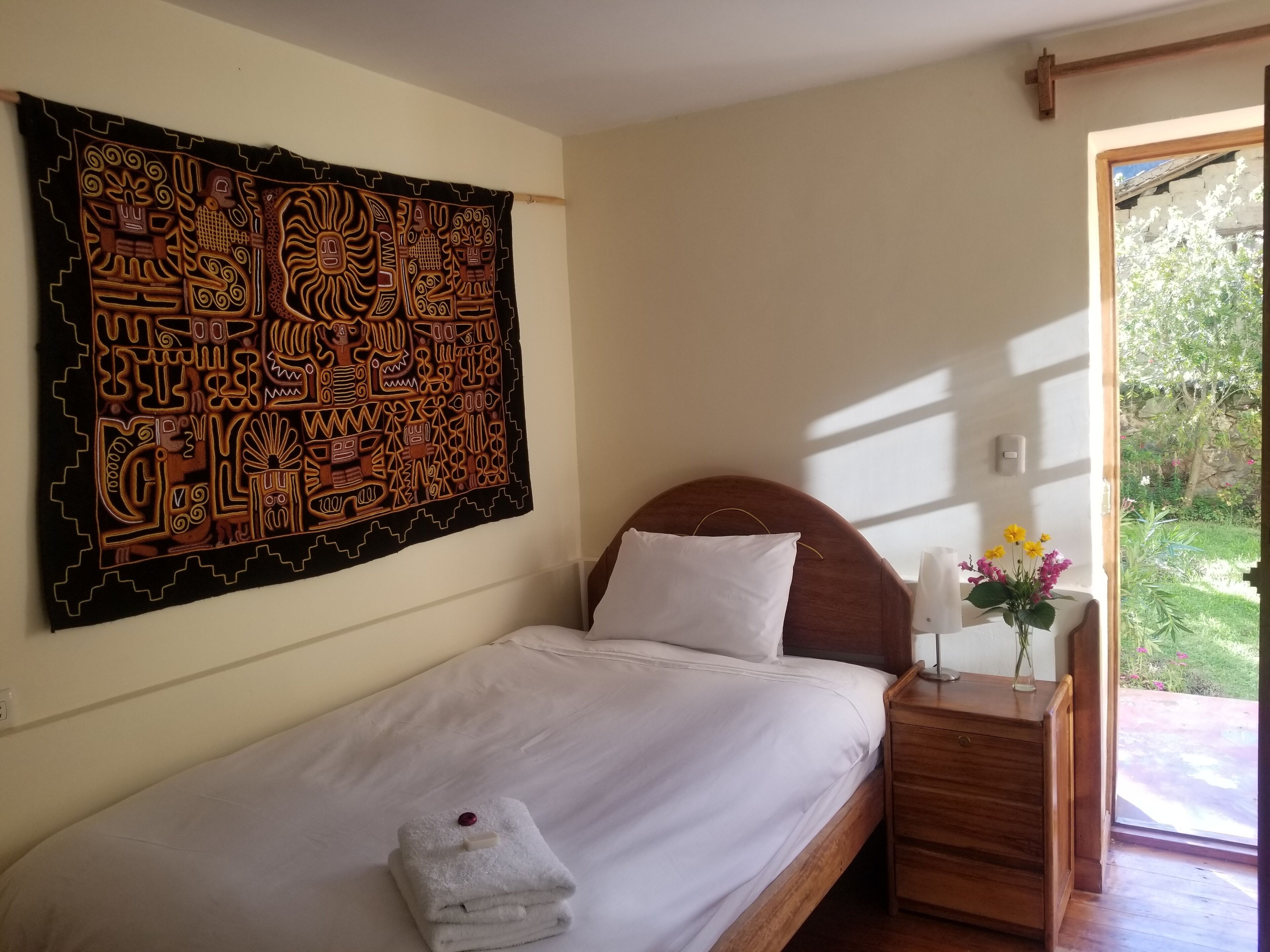 Een eenpersoonsbed met een houten hoofdbord, een wit kussen en een opgevouwen witte handdoek op het bed. Een houten nachtkastje met een bloevas staat naast het bed. Een groot wandtapijt met ingewikkelde motieven hangt aan de muur boven het bed.