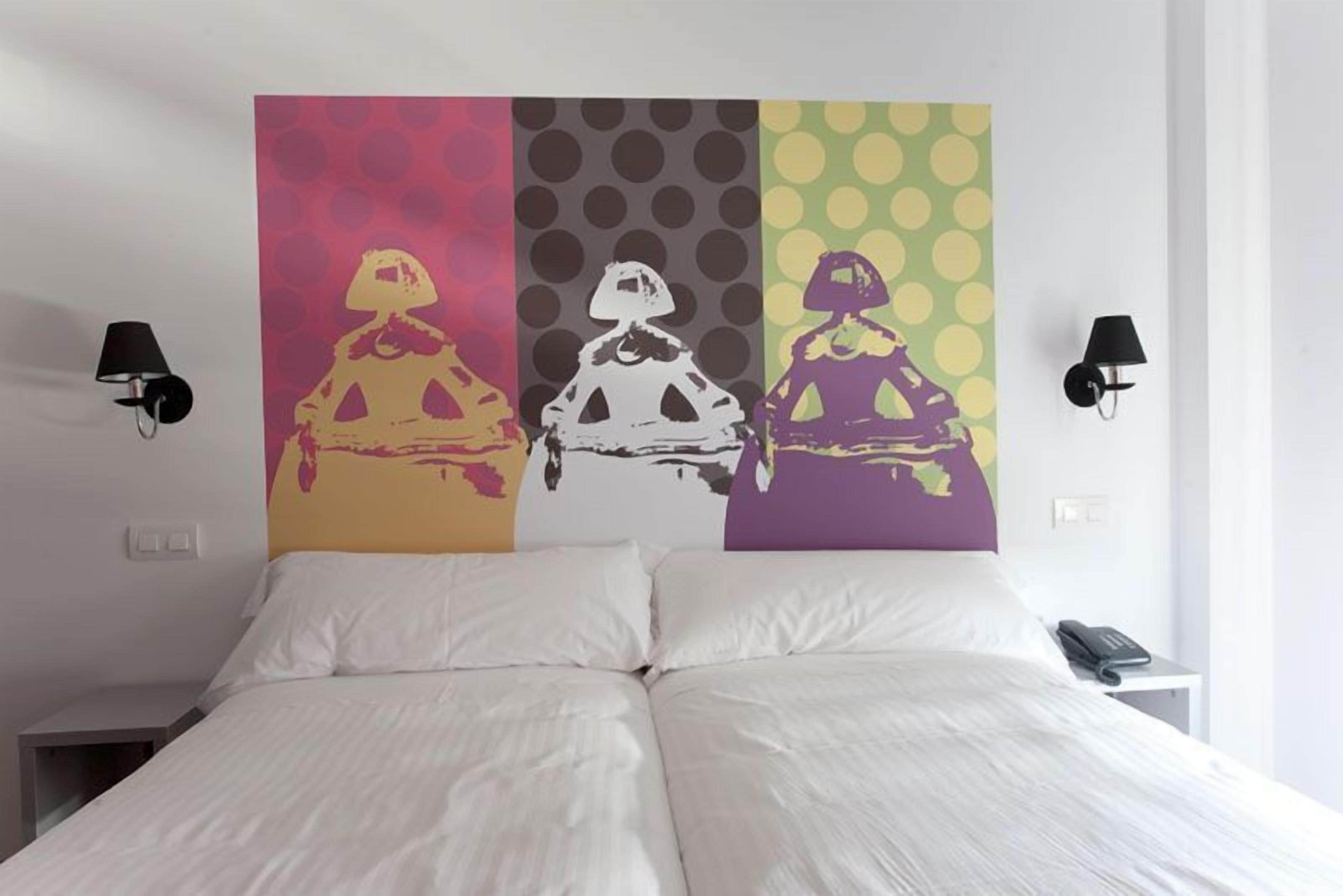 Una cama con ropa de cama blanca, una pared con tres paneles de colores y dos lámparas de pared.