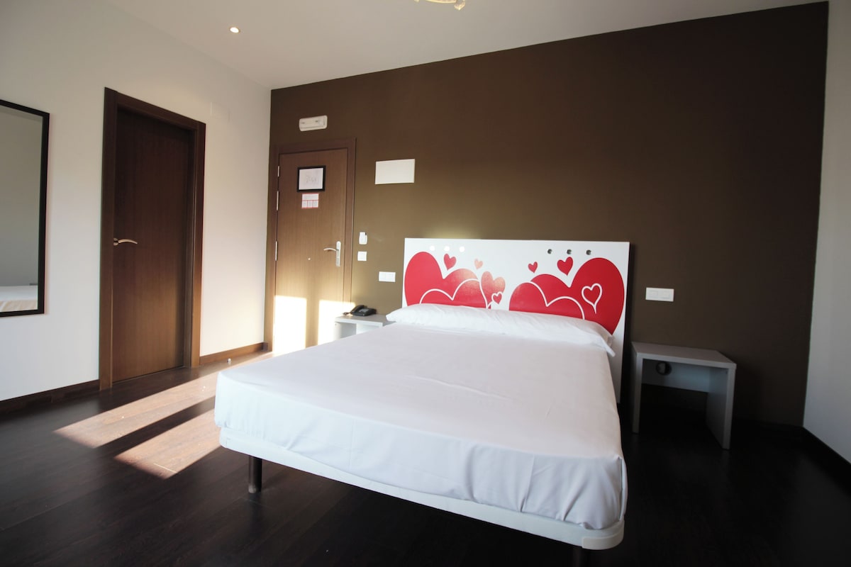 A modern hotel room with a large bed, a bedside table, a nightstand, and a door.