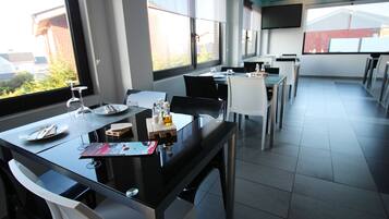 Un comedor moderno con mesas y sillas de vidrio, un televisor de pantalla plana en la pared y amplias ventanas con vistas al exterior.
