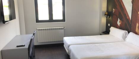 A hotel room with a bed, a desk, a chair, a radiator, a window, and a painting on the wall.