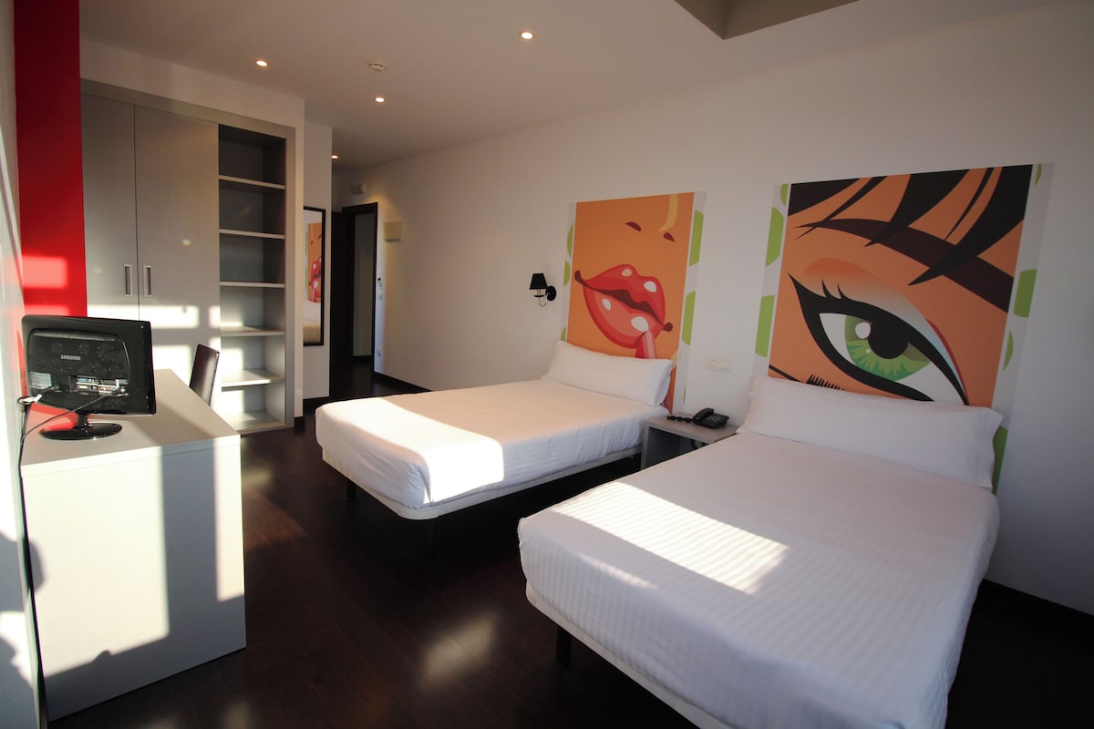 A hotel room with two single beds, a desk with a computer, a wardrobe, and a door leading to another room.
