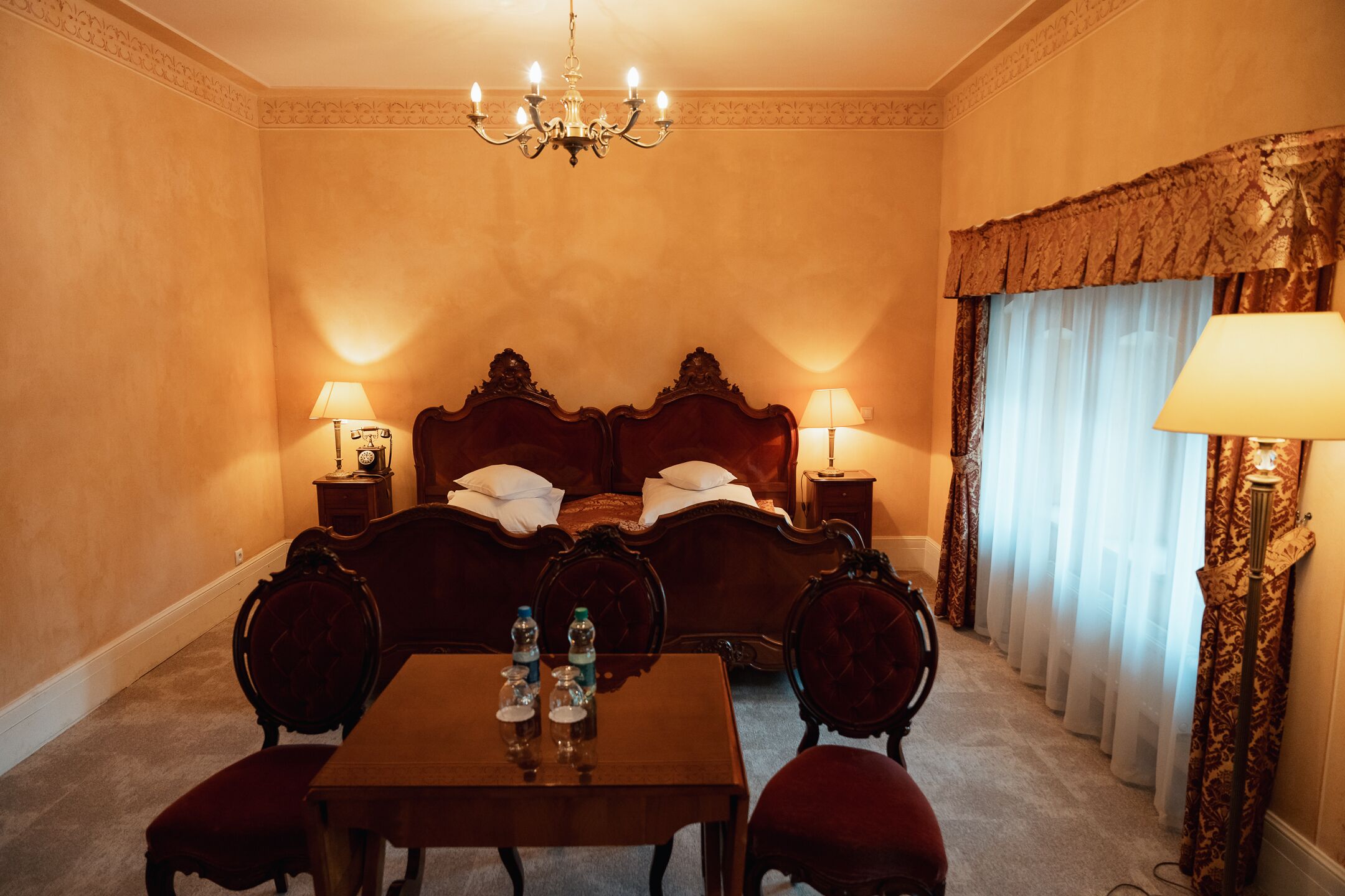 Habitación de hotel con una cama grande, dos mesitas de noche con lámparas, una mesa de madera con sillas y un candelabro.
