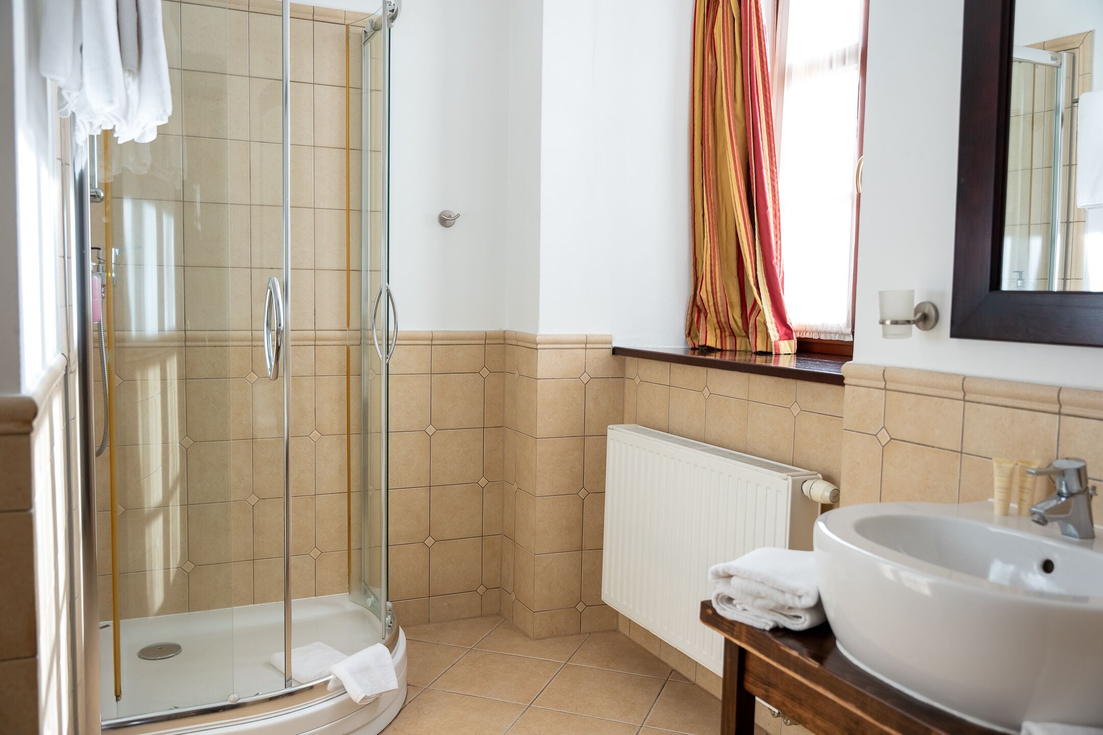 A bathroom with a glass shower enclosure, a white sink with a mirror above it, and a window with curtains.