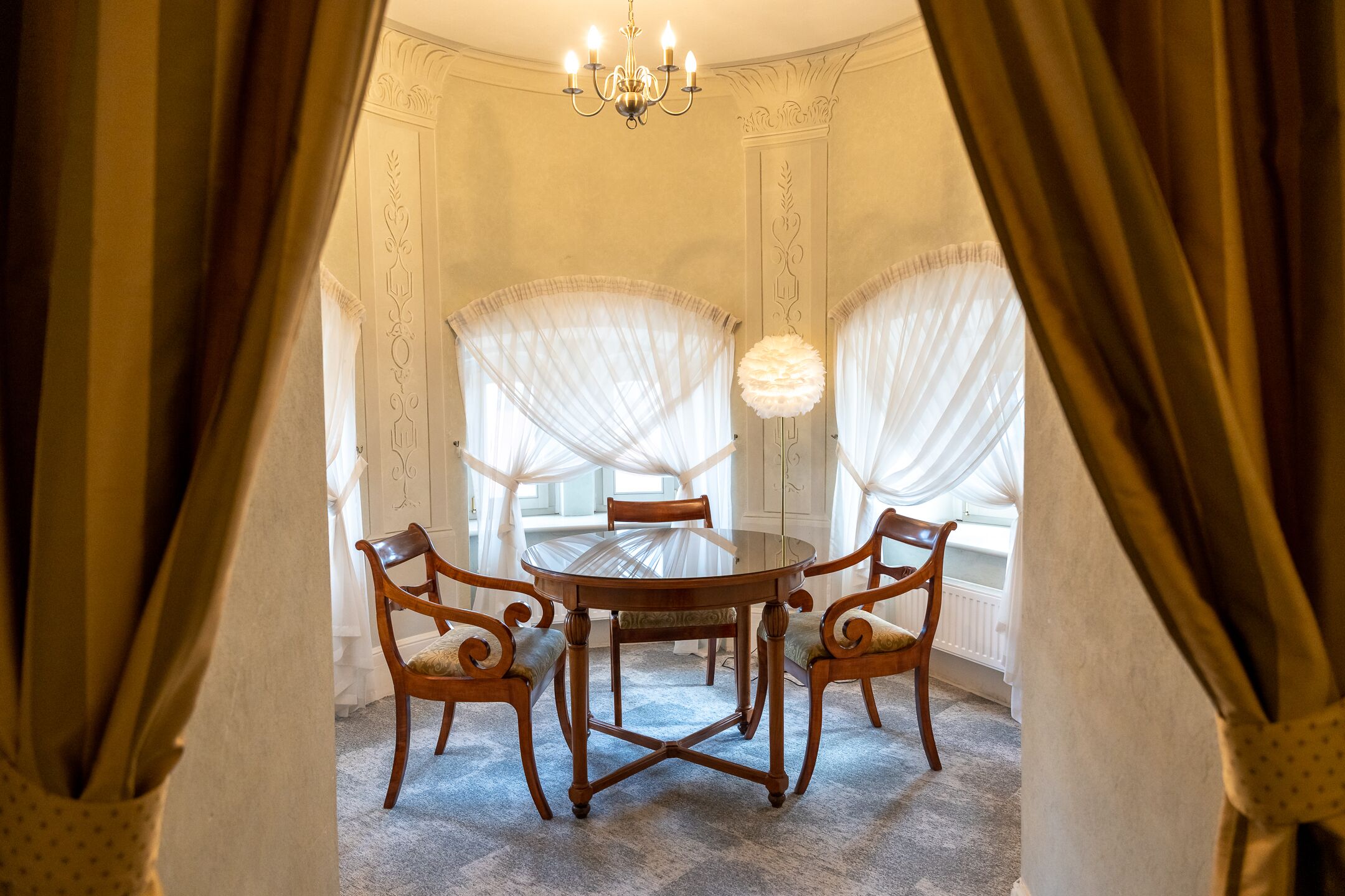 Comedor con una mesa redonda y cuatro sillas, ventanas amplias con cortinas translúcidas y un candelabro.