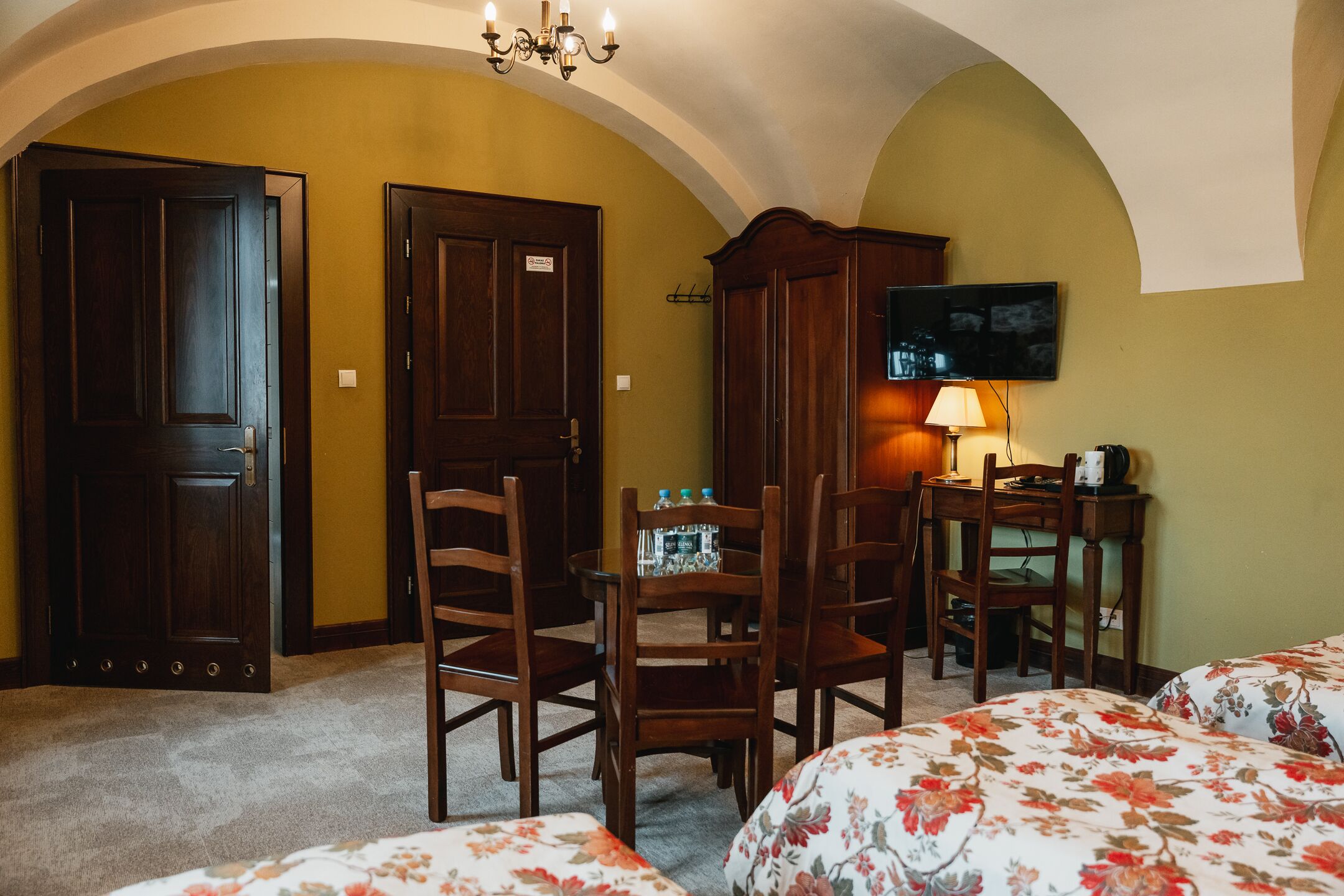 Une chambre d’hôtel avec deux portes en bois, une table à manger avec des chaises, un téléviseur fixé au mur et une literie à motifs floraux.