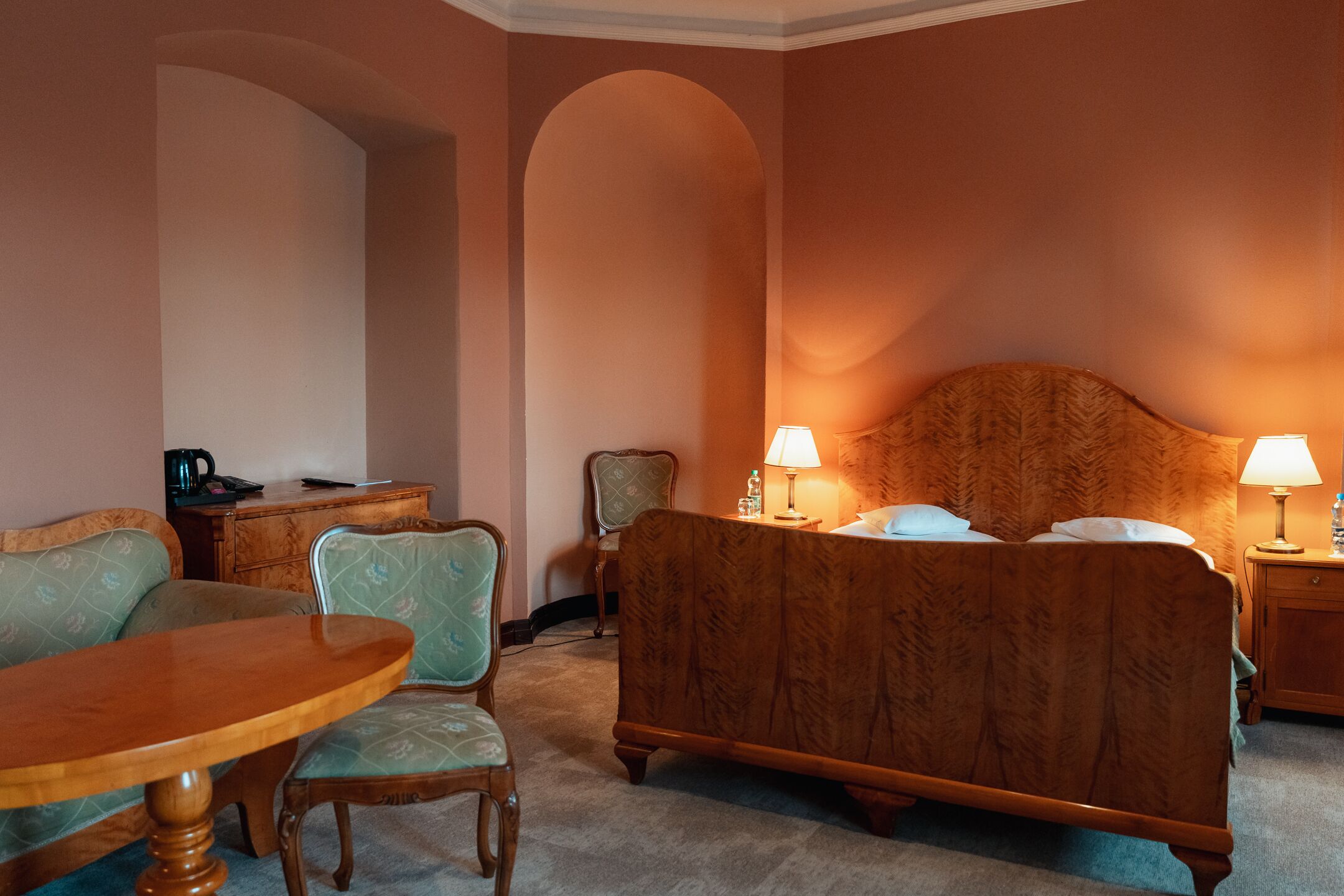 Une chambre d’hôtel avec un lit en bois, deux chaises, une petite table et une table de chevet avec des lampes.