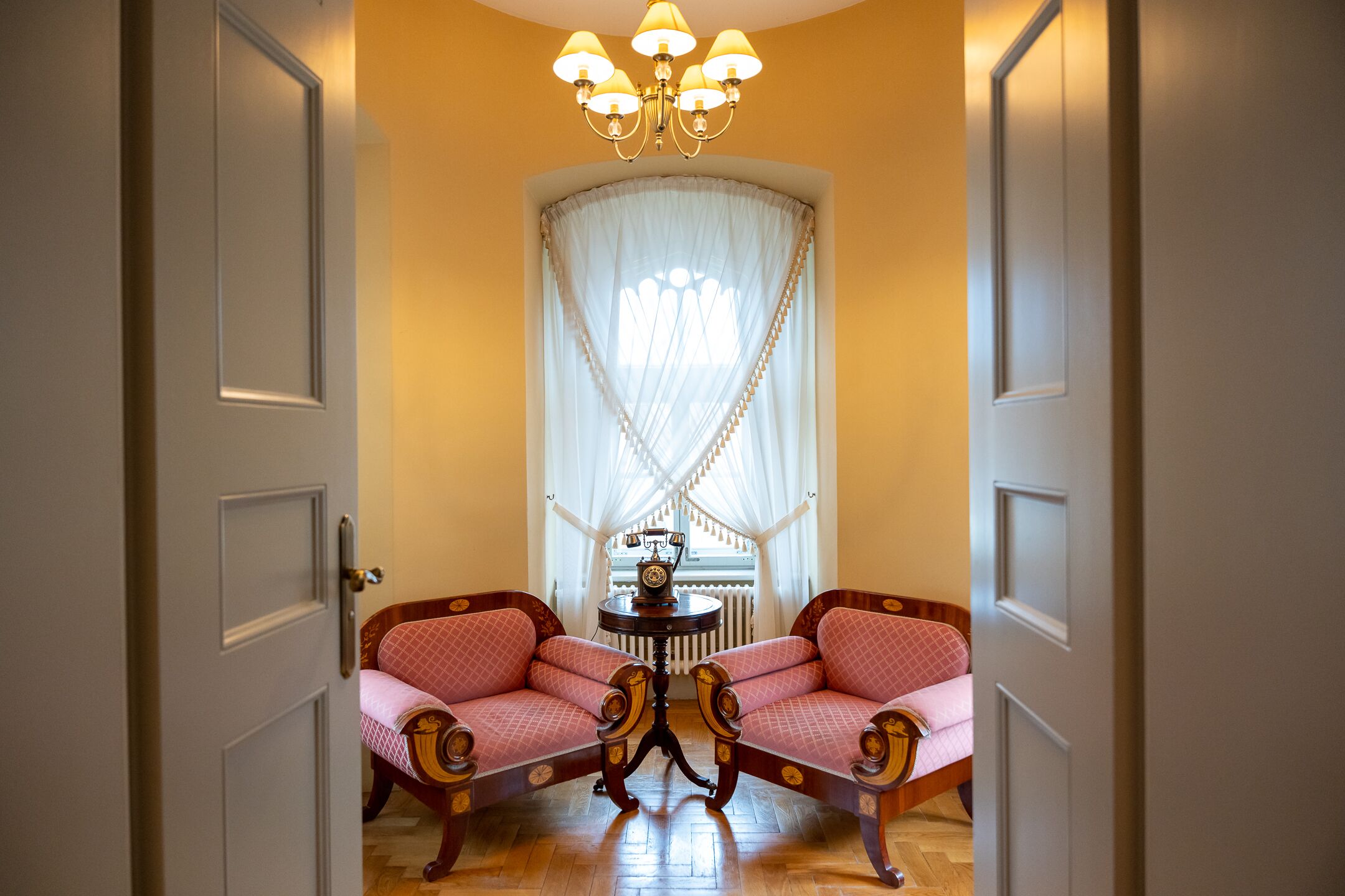 A room with two pink upholstered chairs, a small round table with a clock, and a white-framed window with sheer curtains.