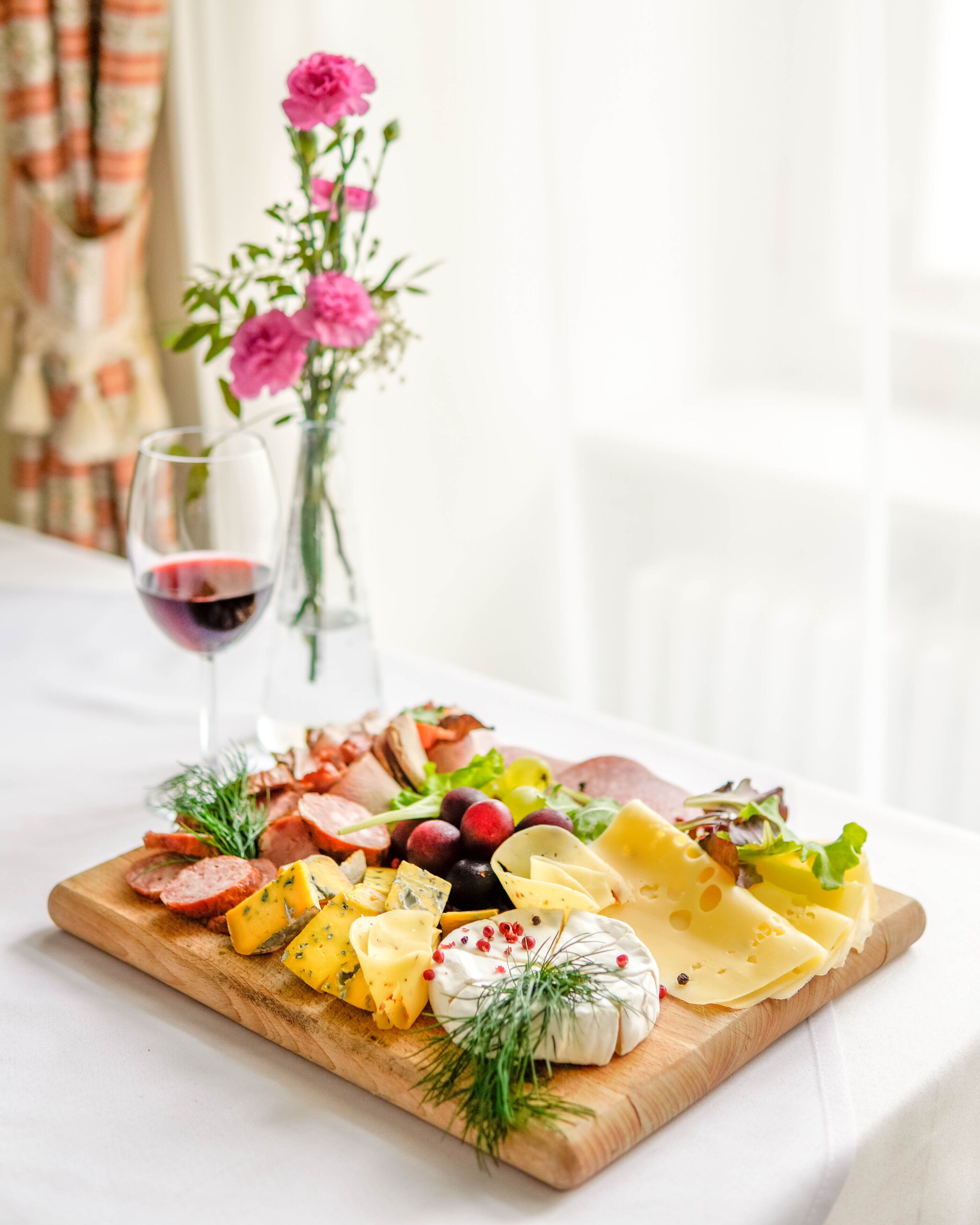 Una tabla de embutidos con diversos quesos, carnes y frutas sobre una tabla de madera, acompañada de una copa de vino tinto y un jarrón de flores.