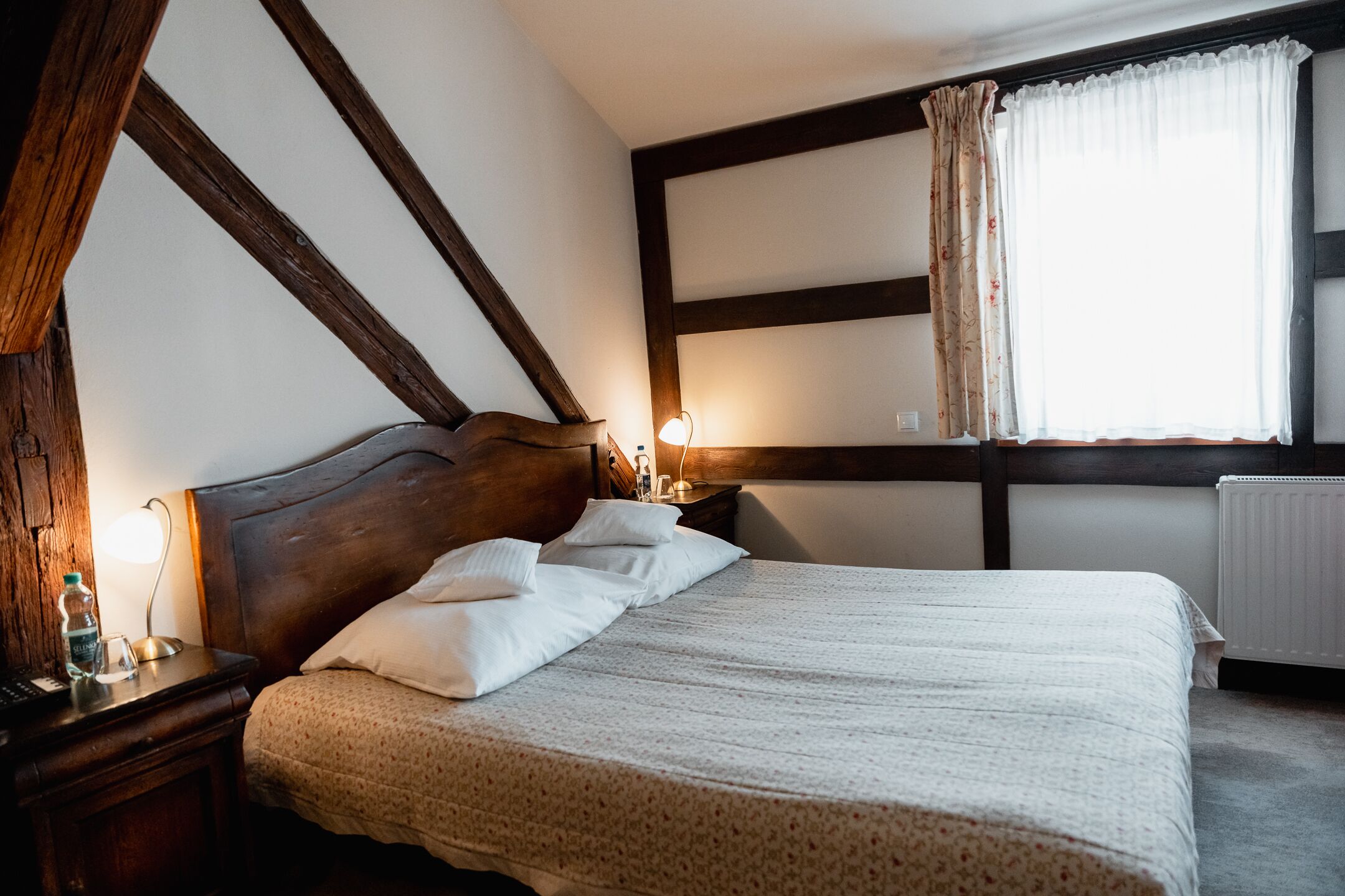 A bedroom with a wooden headboard, a bed with a patterned bedspread, a bedside table with a lamp, and a window with curtains.
