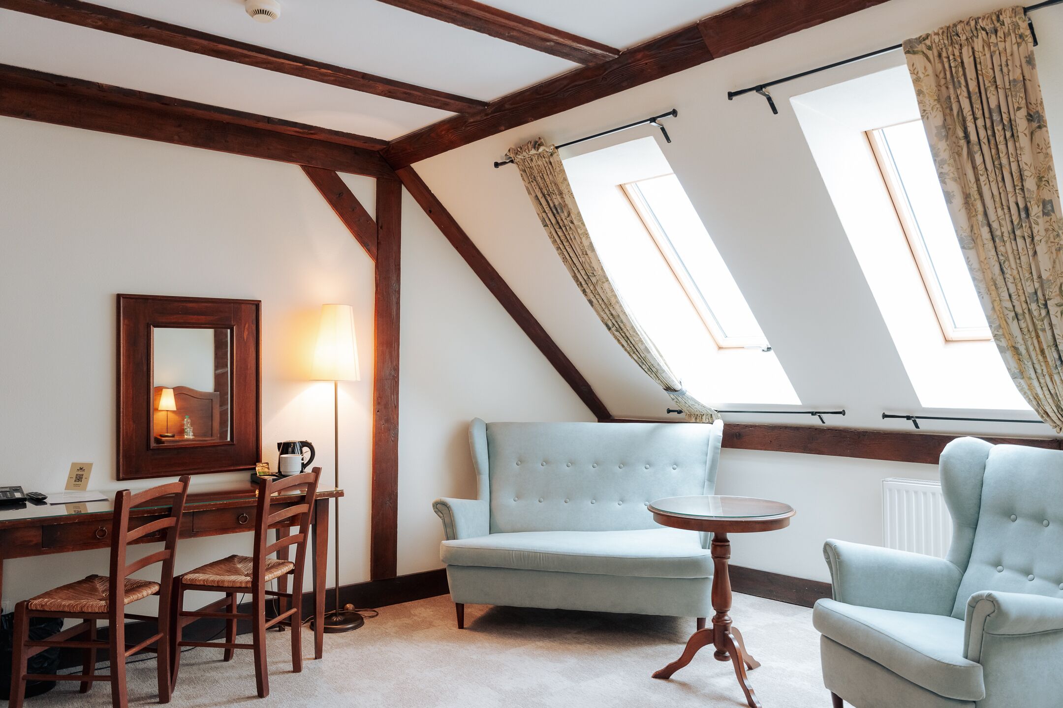 Ein Zimmer mit einem Holztisch, zwei Stühlen, einem Sofa, einem kleinen runden Tisch und einem Spiegel. Zwei Fenster mit Vorhängen und eine schräge Decke mit freiliegenden Balken.