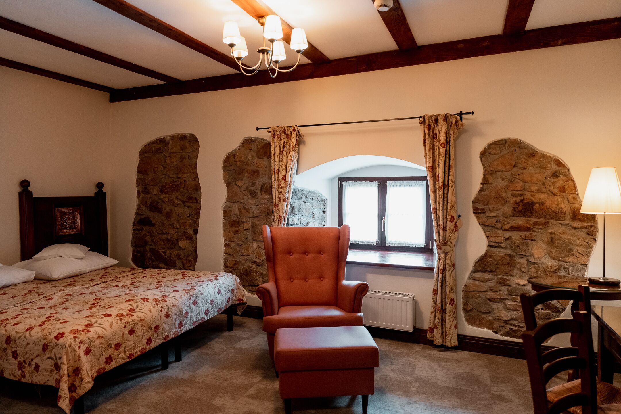 A bedroom with a bed, an orange armchair, a wooden headboard, a stone wall, a window with curtains, and a table with a lamp.