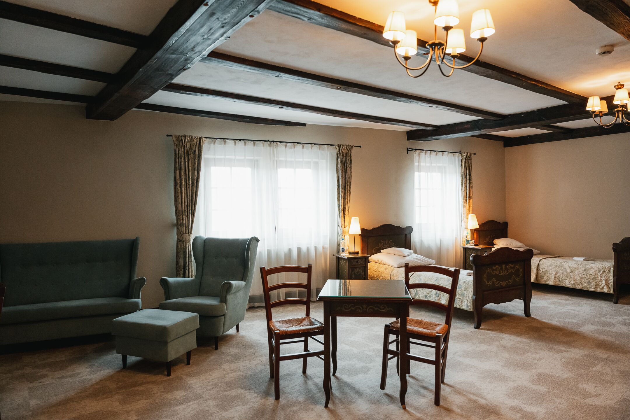 Una habitación con sofá, sillones, mesa y cama. Cortinas cubren las ventanas y un candelabro cuelga del techo.