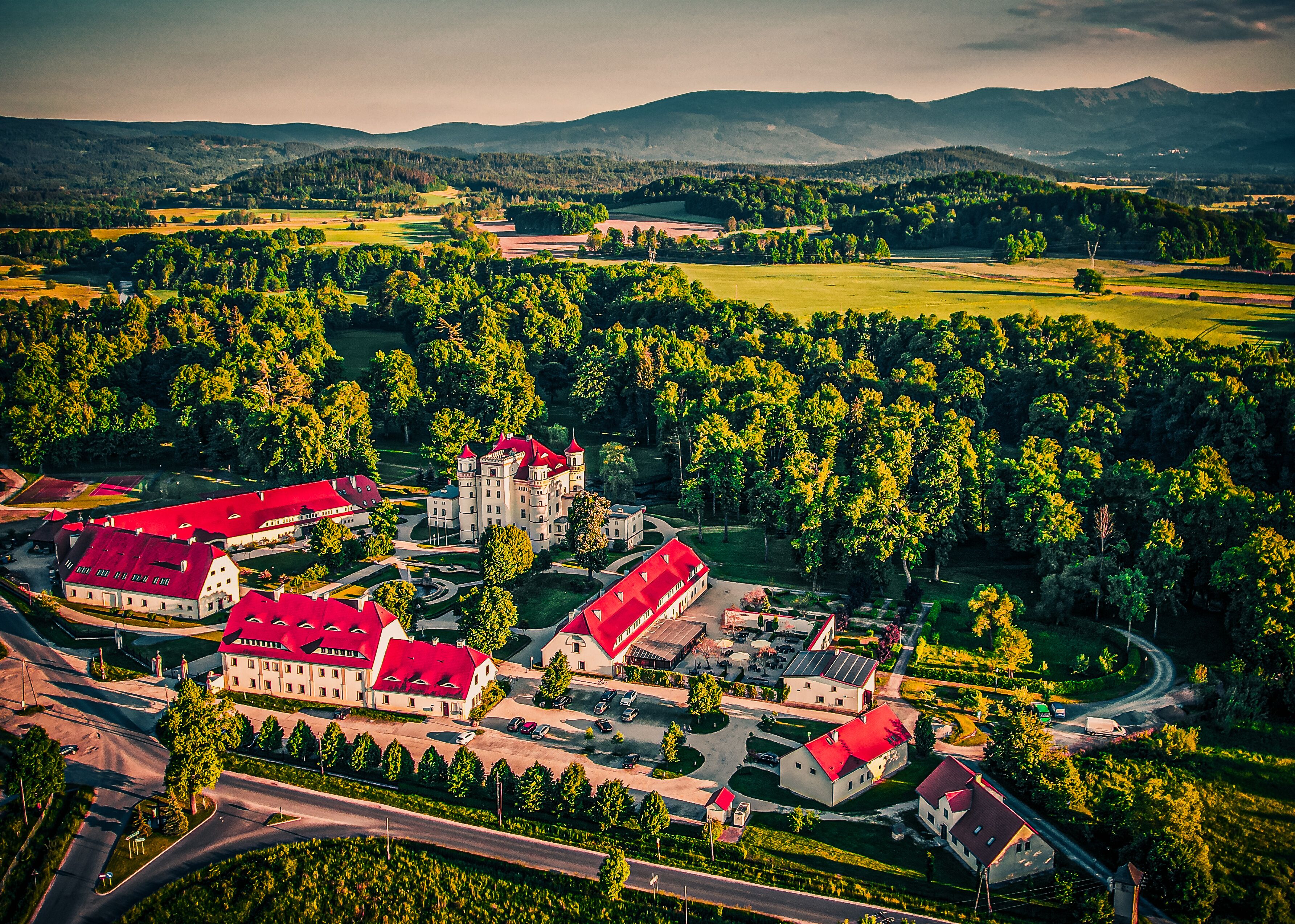An aerial view of a castle complex with multiple buildings, including a central tower, surrounded by lush greenery and rolling hills in the background.