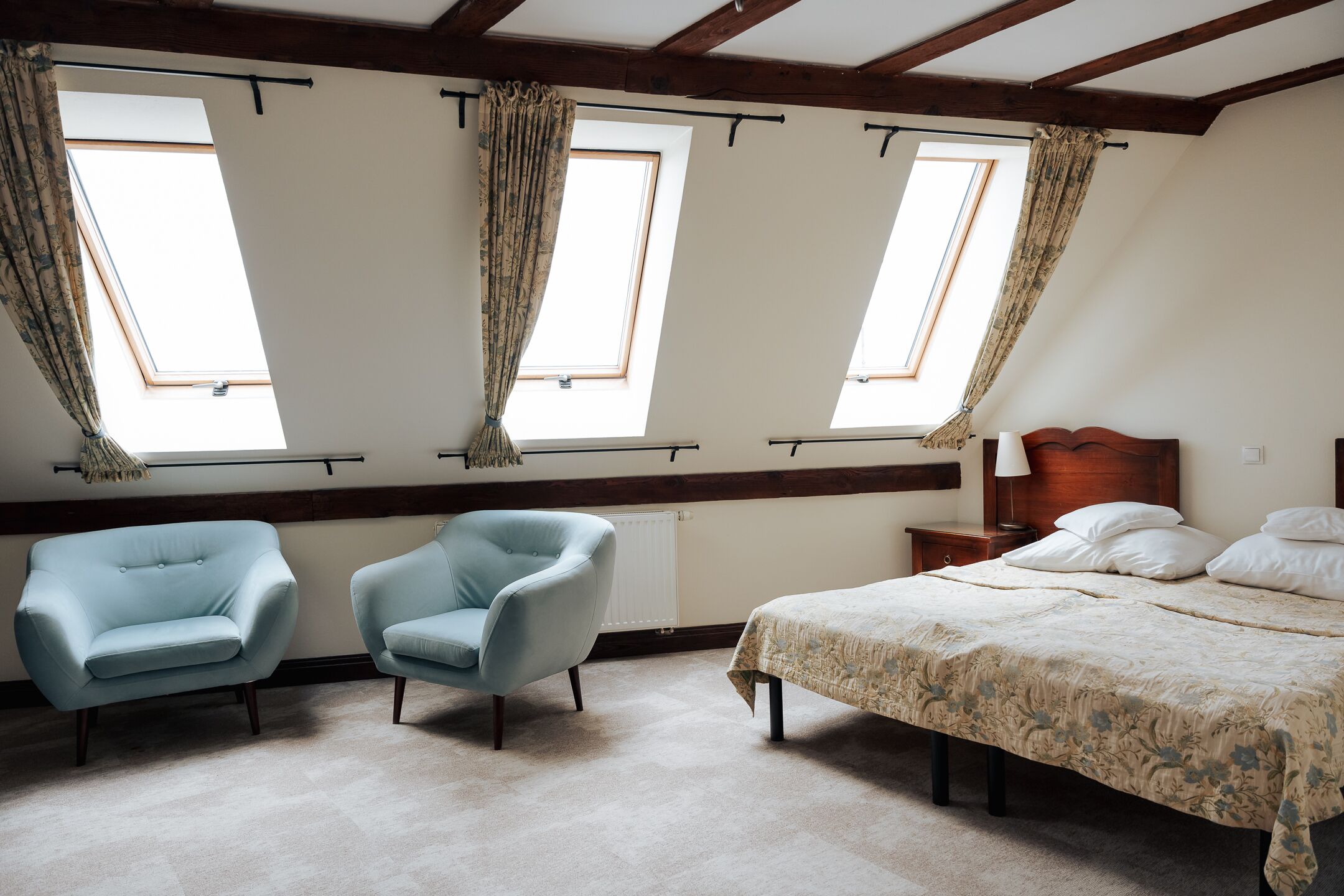 Ein Schlafzimmer mit einem großen Bett, zwei Sesseln und drei Fenstern mit Vorhängen.