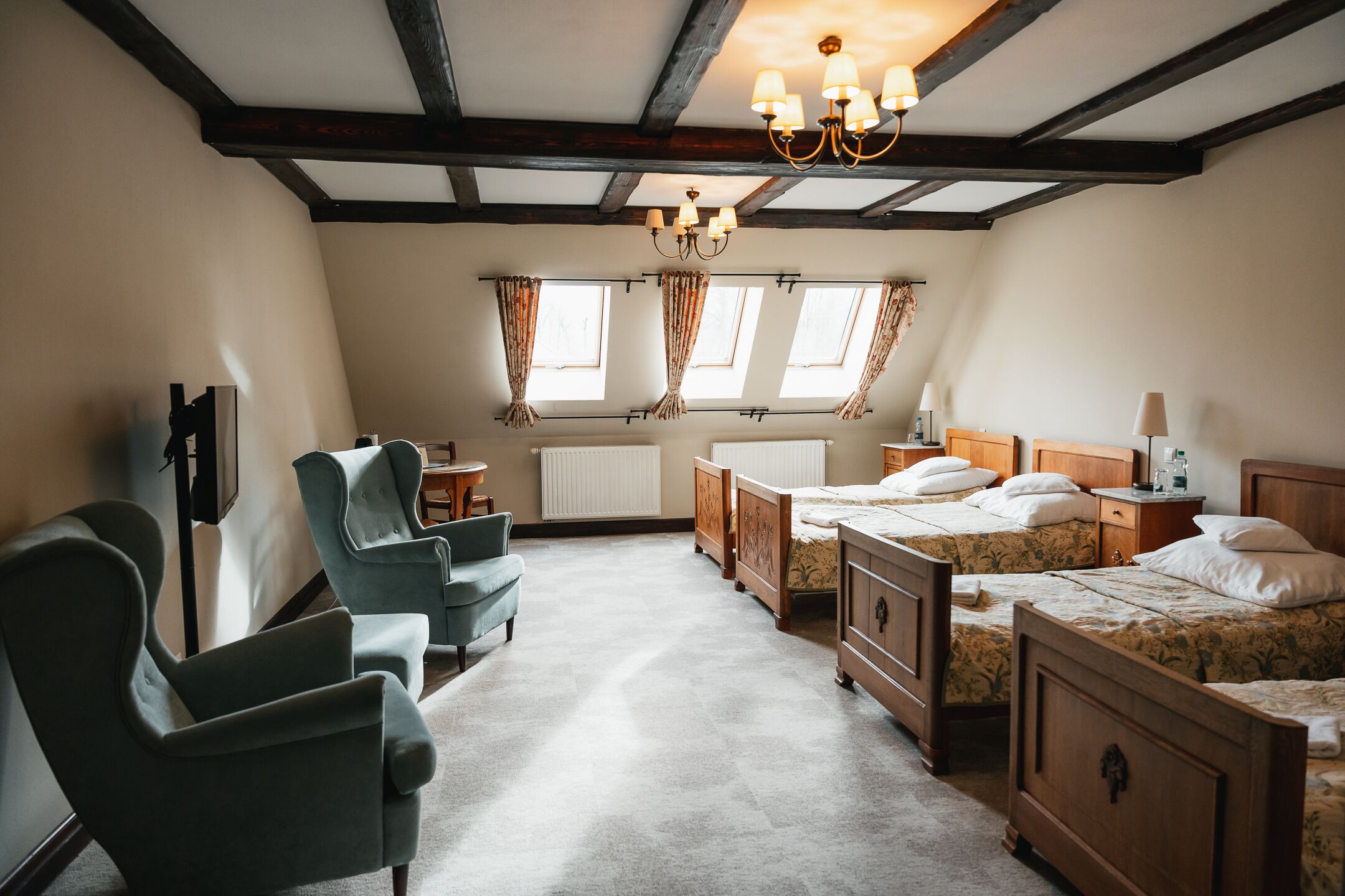 Une chambre d’hôtel avec un plafond haut, des poutres apparentes, un lustre, une grande fenêtre avec des rideaux, un radiateur et un lit doté d’une tête de lit en bois.