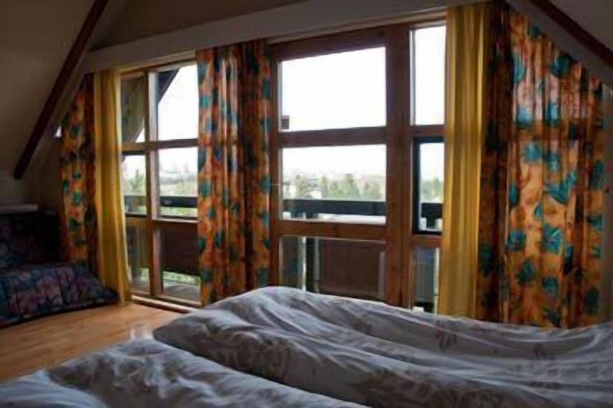 A bedroom with a large bed, a view of the outdoors through a window, and a balcony with curtains.