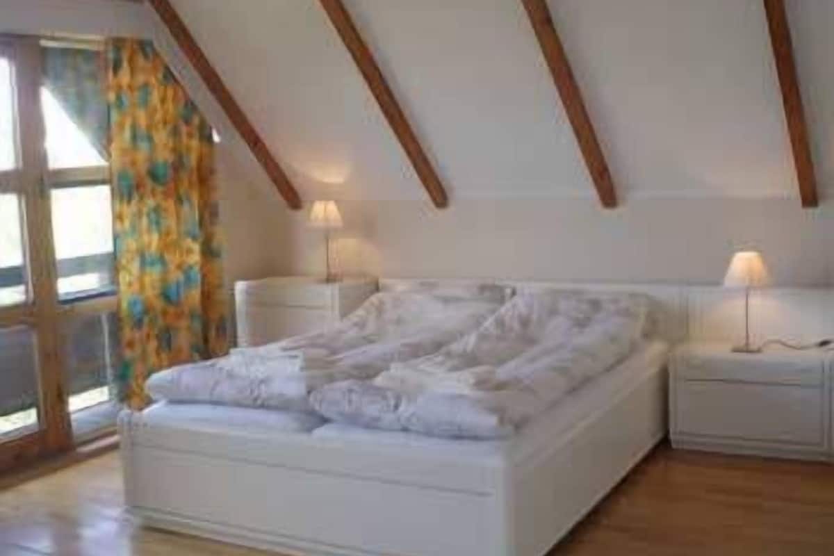 A bedroom with a large bed, wooden beams on the ceiling, two bedside tables with lamps, and a window with curtains.